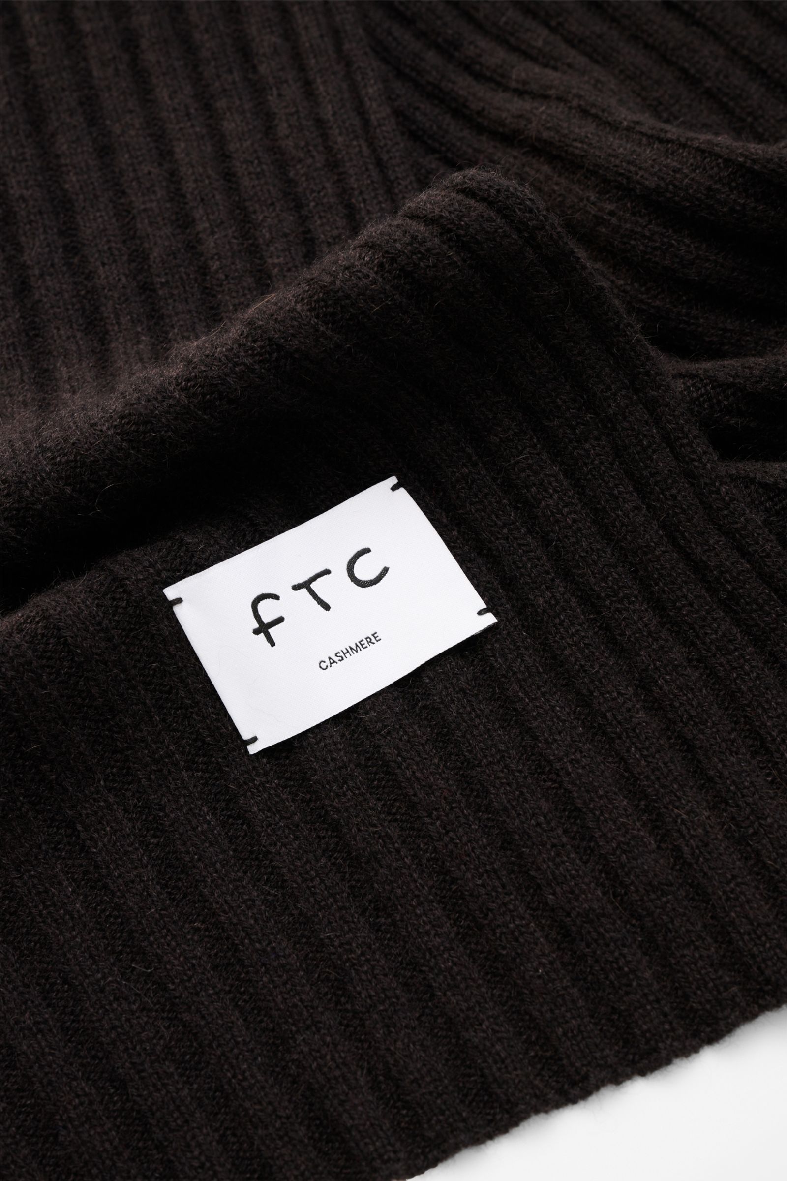 FTC Cashmere Schal dunkelbraun mit breitem Rippstrickmuster, weichem Griff und großem Logo-Weblabel, Nahaufnahme von oben.