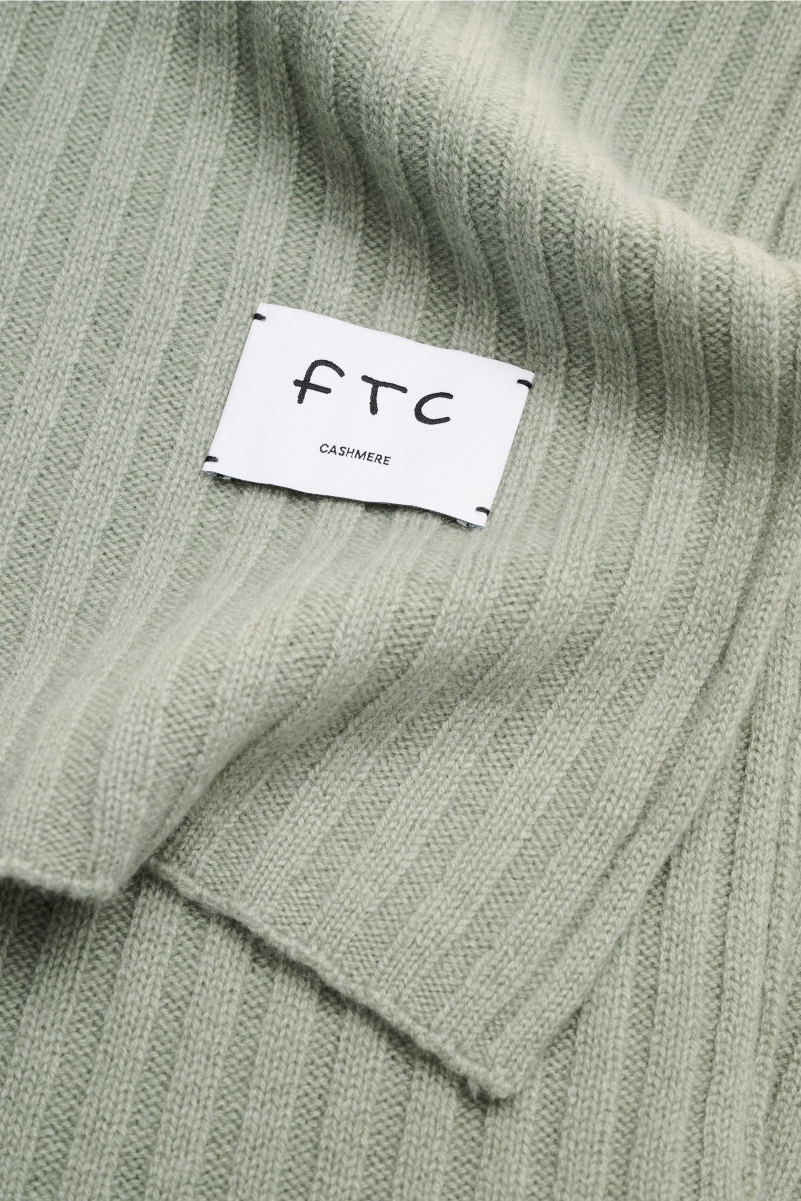 Nahaufnahme des FTC Cashmere Schal graugrün mit breitem Rippstrickmuster, weichem Griff und großem Weblabel, von oben fotografiert.