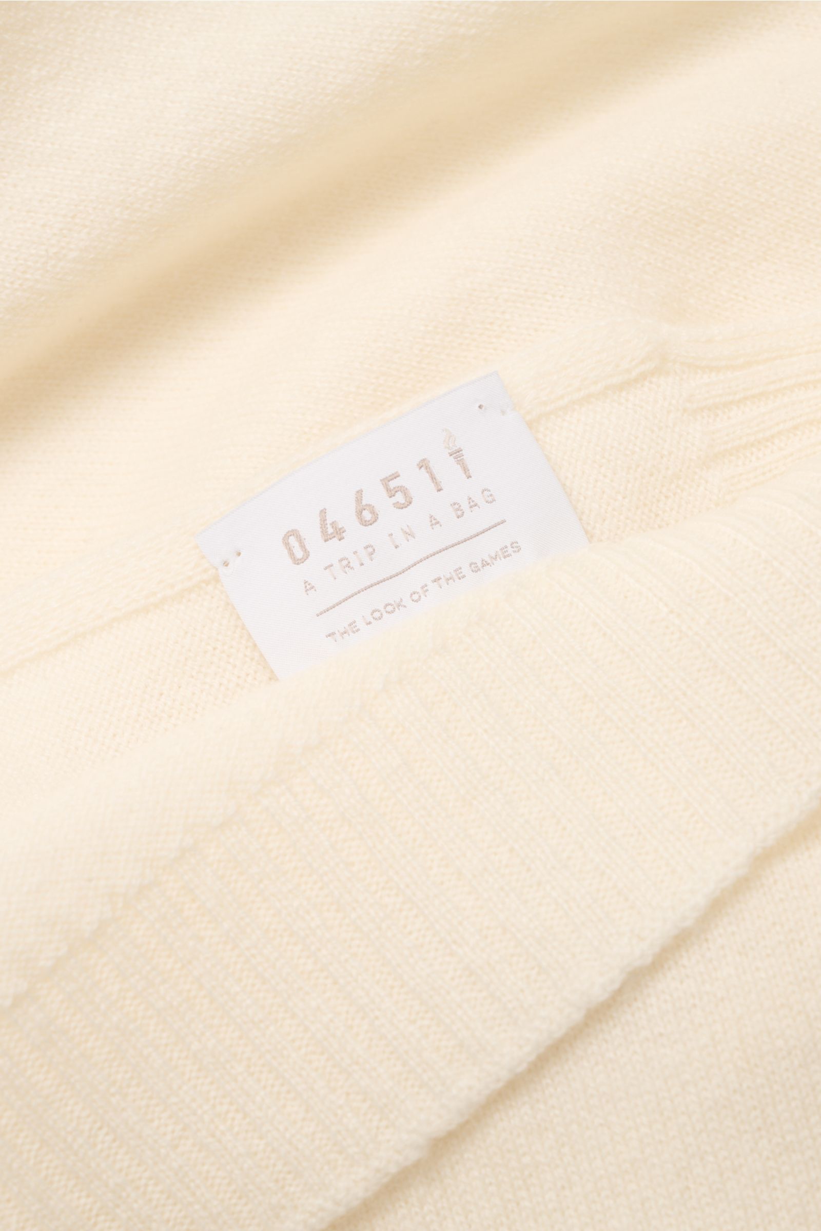 Nahaufnahme aus schräger Vogelperspektive eines cremefarbenen 04651/ A trip in a bag Cashmere Schals mit weichem Griff, Rippstrick-Kanten und Etikett.