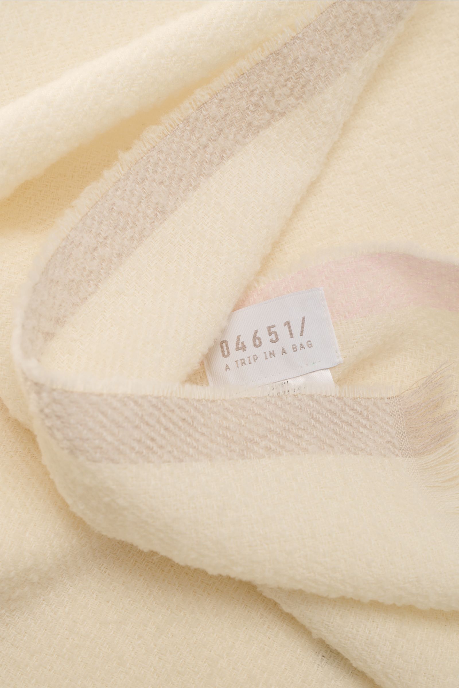 Close-up top-down view of the 04651/ A trip in a bag Scarf cream showing its fine virgin wool bouclé texture, narrow fringed edge, and label. Luxurious comfort for winter: Fine virgin wool material makes this scarf in bouclé look from 04651/ A TRIP IN A B