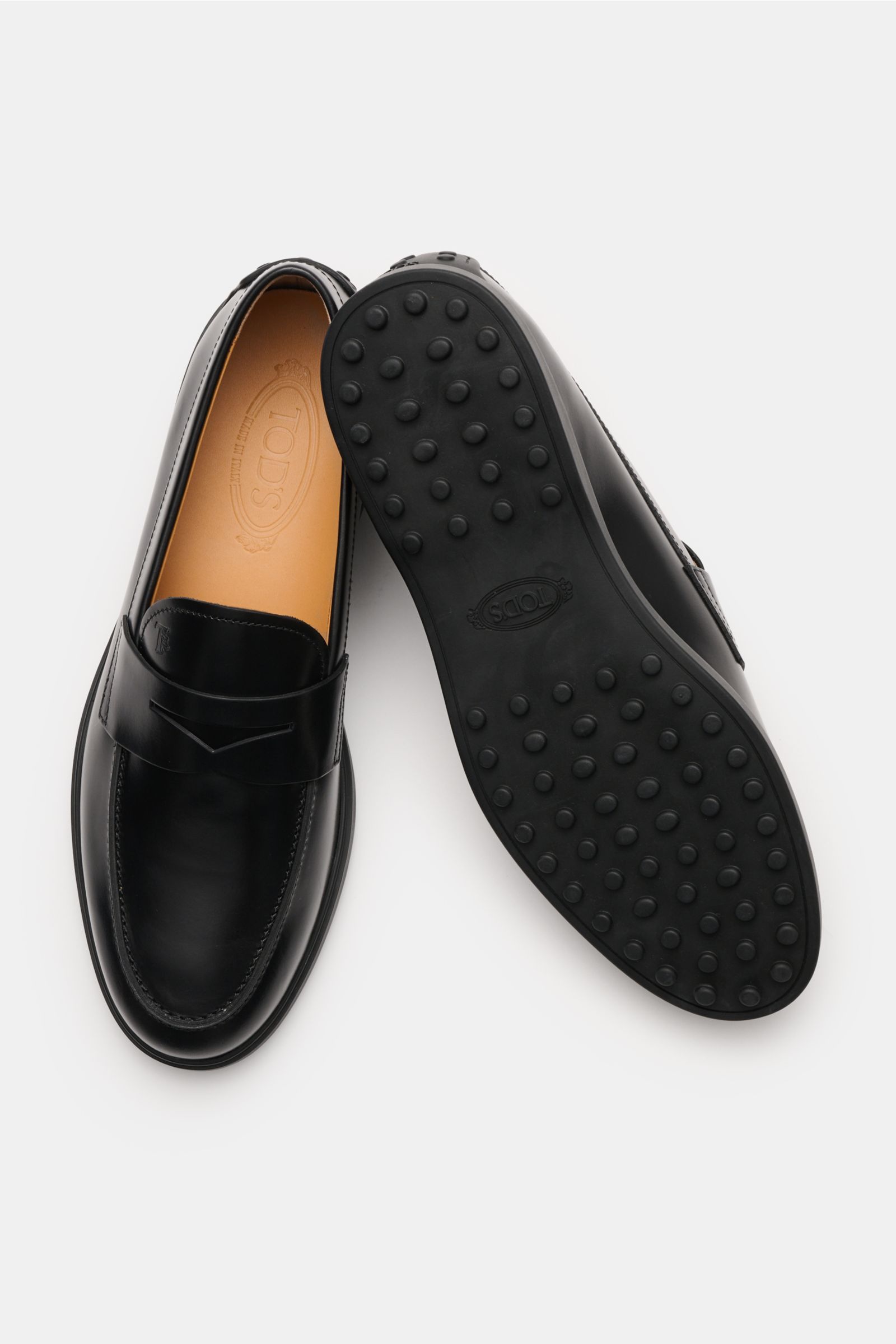 Tod's Penny Loafer schwarz, aus edlem Glattleder, leicht schmal zulaufend, mit typischem Penny Strap, Steppnaht auf der Vorderkappe, Noppendetail an der Ferse, Laufsohle aus Gummi mit Noppen, made in Italy, von oben fotografiert, ein Schuh zeigt die Sohle