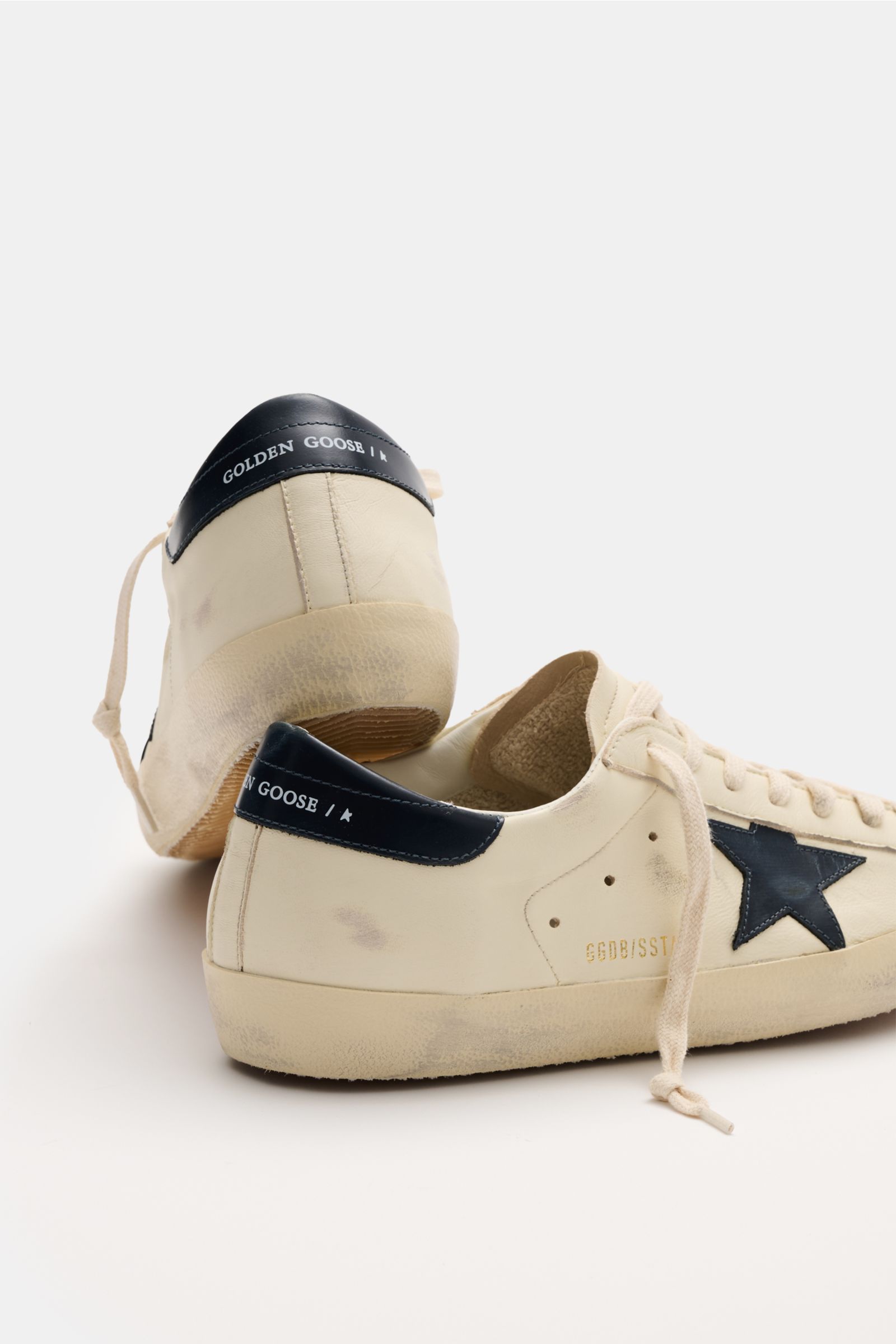 Golden Goose Sneaker 'Super-Star Classic' offwhite/hellgrau shown from a rear and side angle, featuring vintage-finish off-white leather with a black star, black heel tab with logo, seven-hole lacing, padded collar, and worn rubber sole. Für alle, die Pat