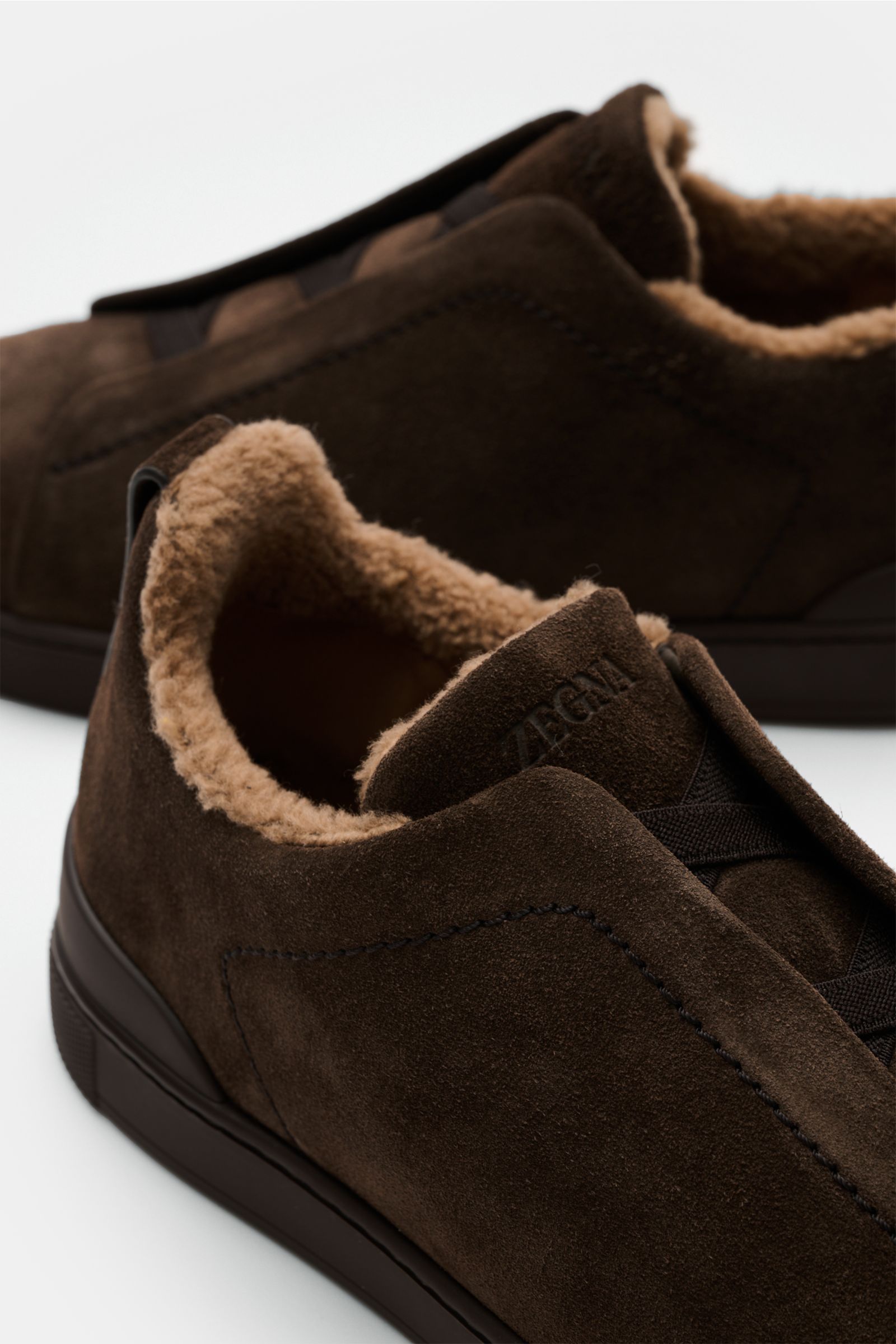 Zegna Slip-on-Sneaker 'Triple Stitch' dunkelbraun aus edlem Veloursleder mit Fellimitatfutter, elastischen Bändern, ZEGNA-Prägung, flexible Gummisohle, Nahaufnahme seitlich oben.