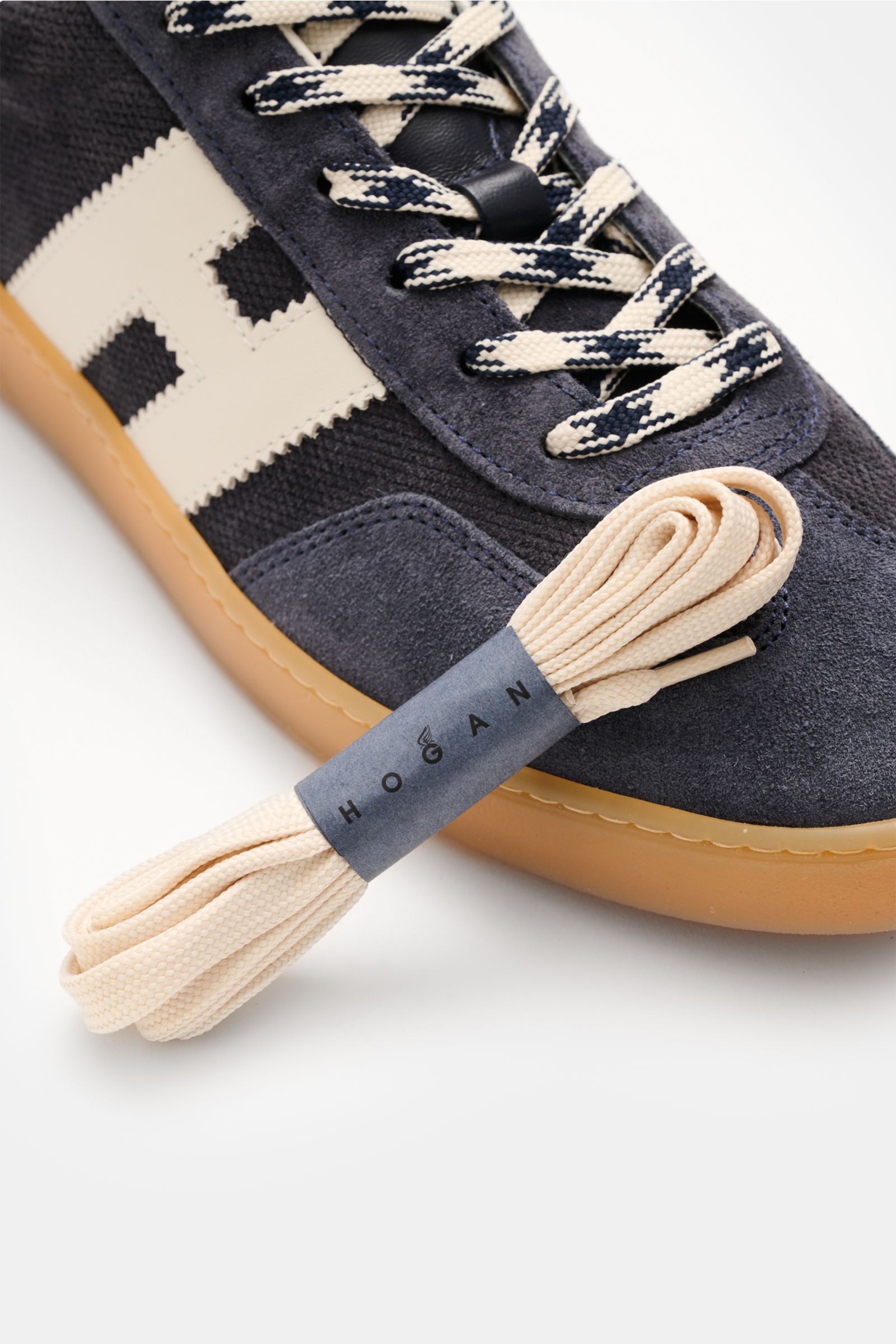 Close-up angled view of Hogan Sneaker 'Allacciato' rauchblau/weiß showing embossed suede, six-hole lacing, logo-printed tongue, padded collar, H-shaped brand initial, leather-textile lining, memory foam insole, and flexible gum sole with extra beige laces