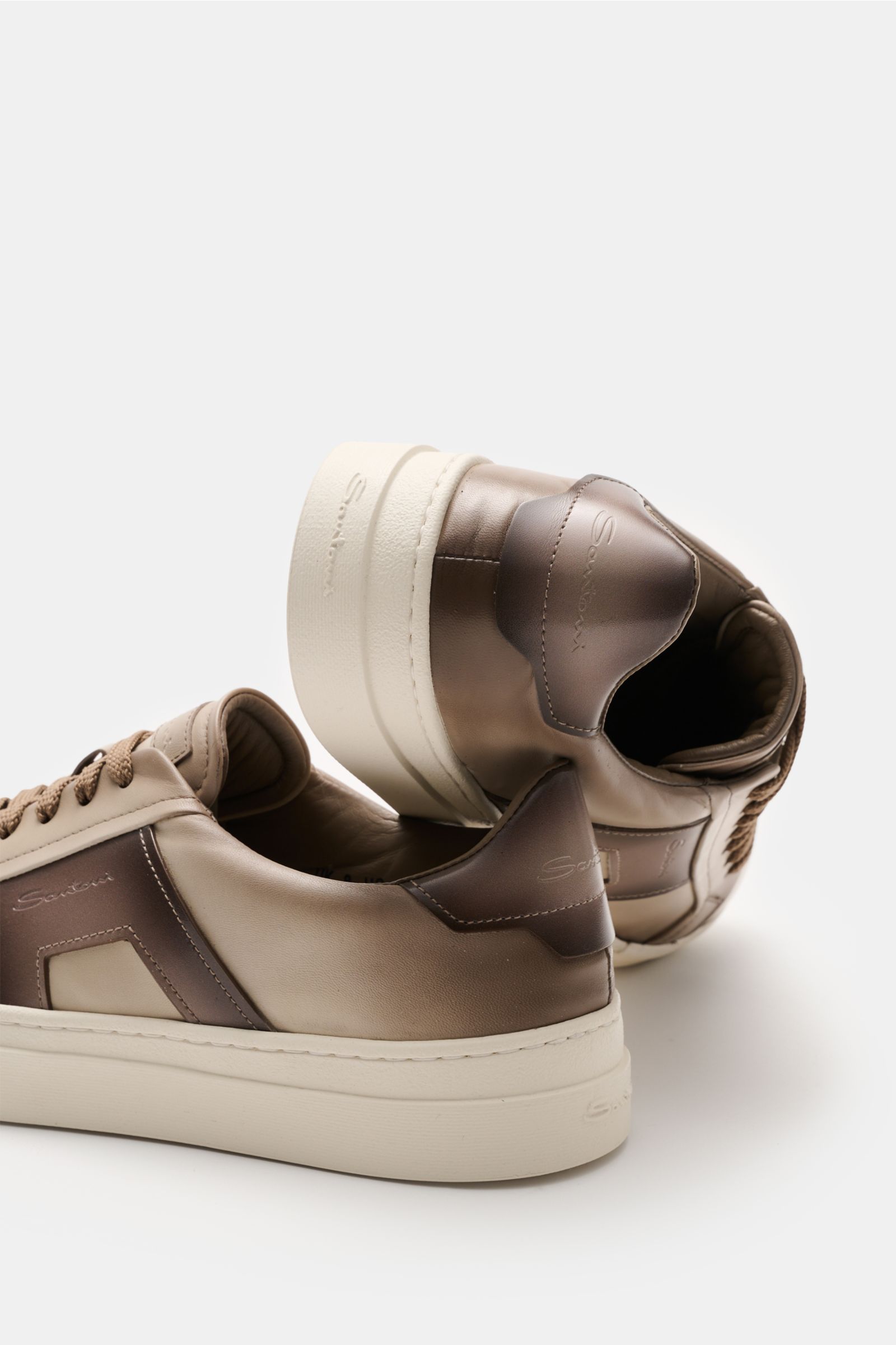 Santoni Sneaker 'Double Buckle' taupe shown from the back and side, featuring fine smooth leather, characteristic double buckle overlays, round shape, seven-eyelet lacing, emblem on the tongue, padded collar, leather lining, and flexible rubber sole. Homm