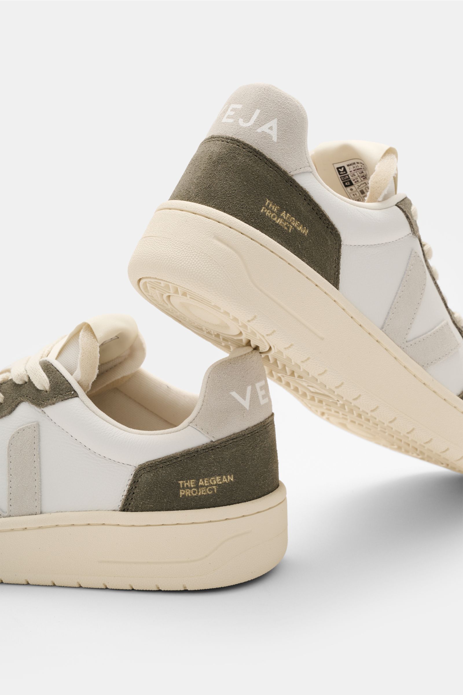 Veja Sneaker 'V-82' offwhite/taupe/hellgrau shown from a low rear-side angle, featuring sustainable leather, round perforated toe, seven-hole lacing, padded collar with logo, suede overlays, contrasting V-overlay, jersey lining, and recycled rubber sole. 