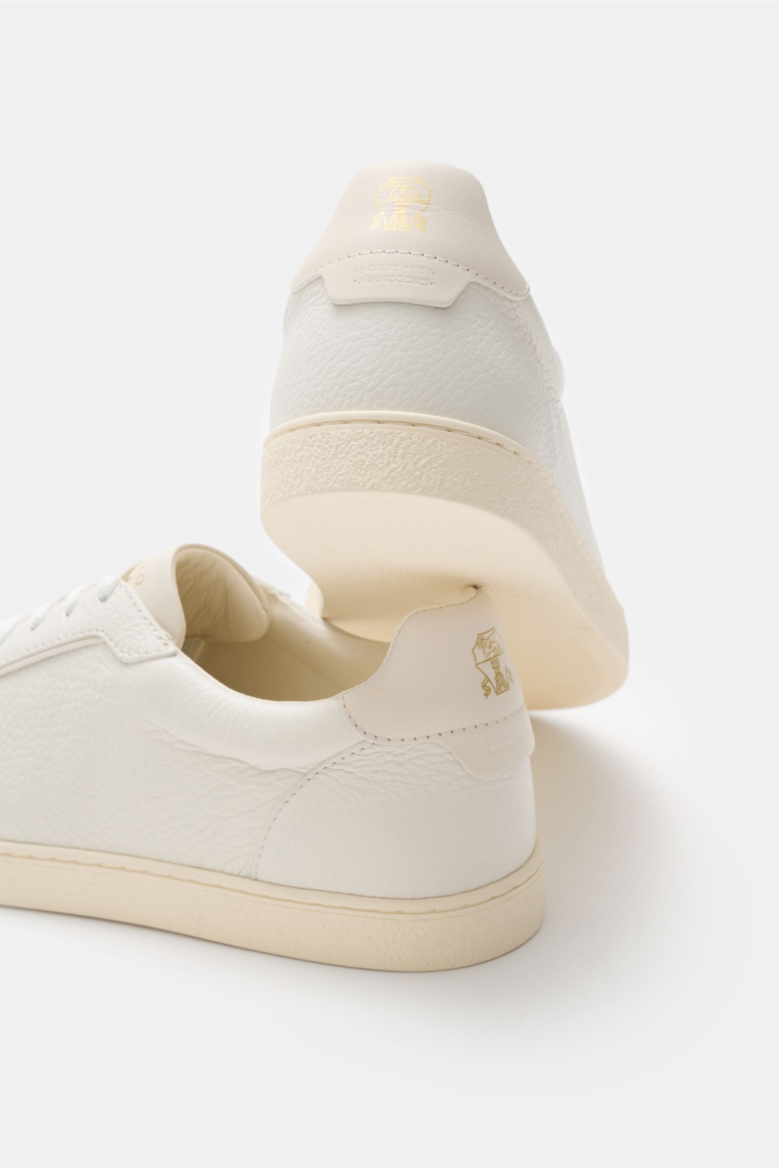 Brunello Cucinelli Sneaker creme aus geschmeidigem Leder, seitlich und von unten fotografiert, retro minimalistisch mit goldfarbener Label-Prägung.
