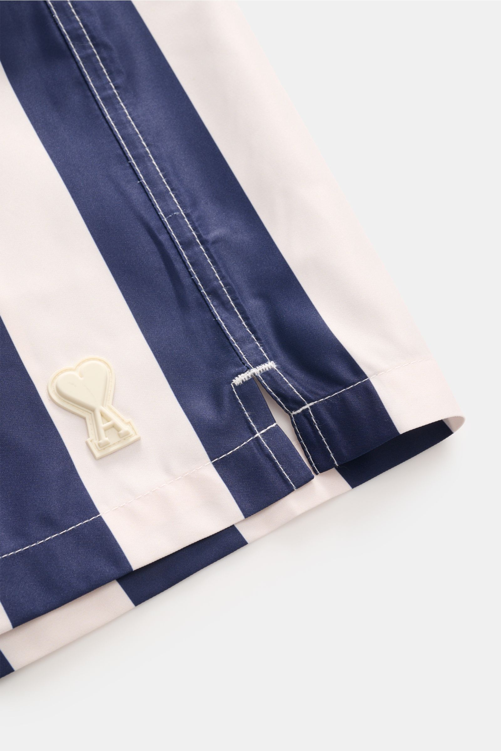 Close-up side view of Ami Paris Badeshorts navy/weiß gestreift showing navy and white stripes, stitching, and rubber logo detail.

Description: Lässig durch die warme Saison: Mit den Badeshorts von AMI PARIS überzeugen Sie mit französischem Look und genie
