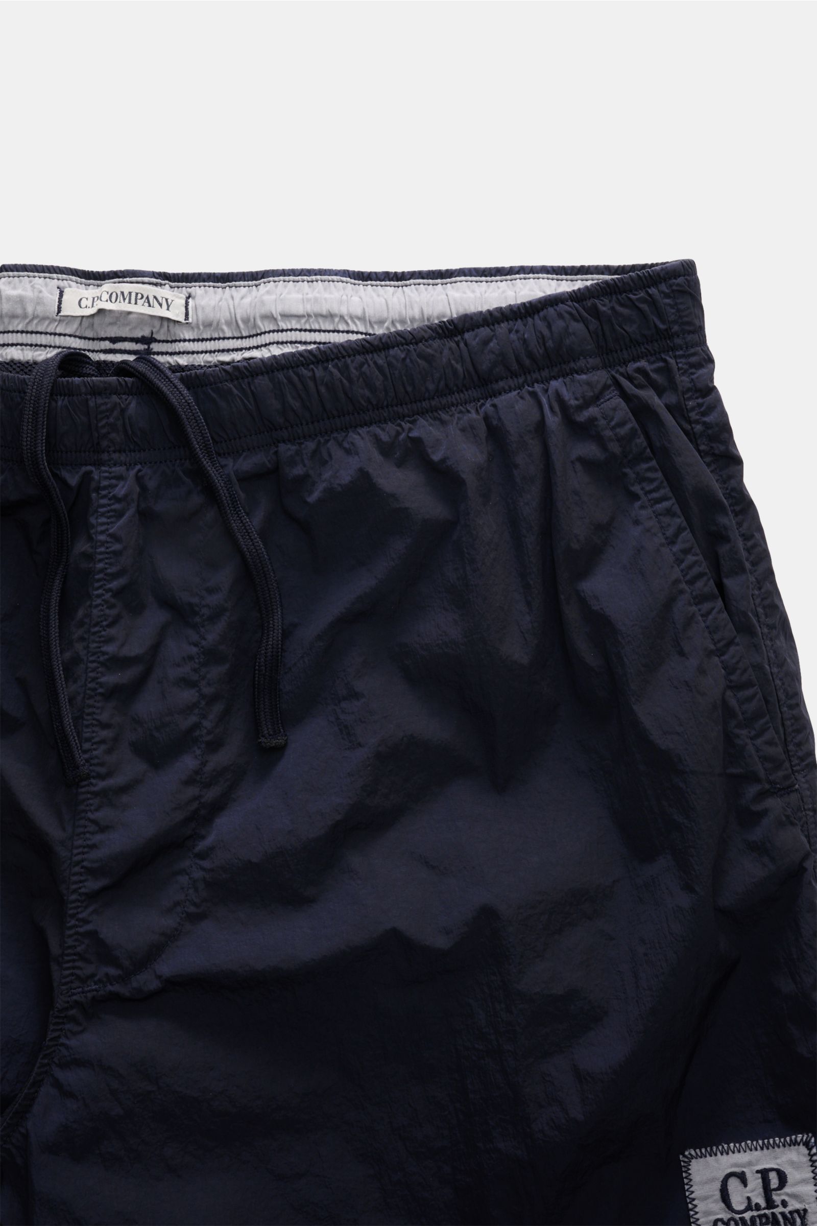 C.P. Company Badeshorts navy mit glänzender Oberfläche, elastischem Bund, Tunnelzug, Mesh-Innenfutter, französischen Taschen, Logo-Patch, fotografiert aus Frontperspektive.