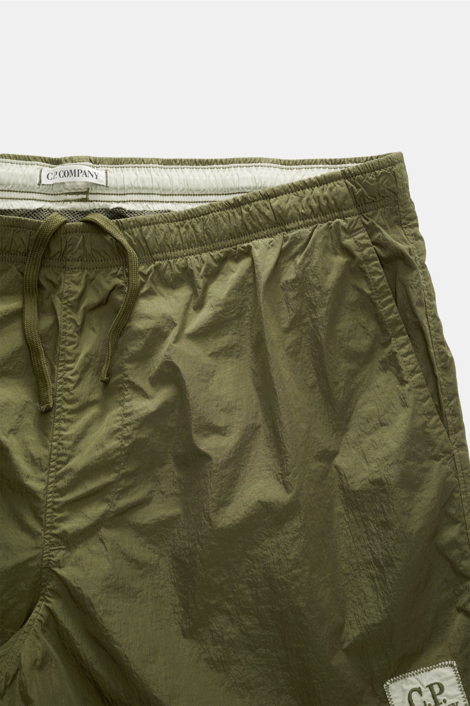 C.P. Company Badeshorts oliv, Nahaufnahme von oben, elastischer Bund mit Tunnelzug, glänzende Oberfläche, französische Taschen, Mesh-Innenfutter, Regular Fit.