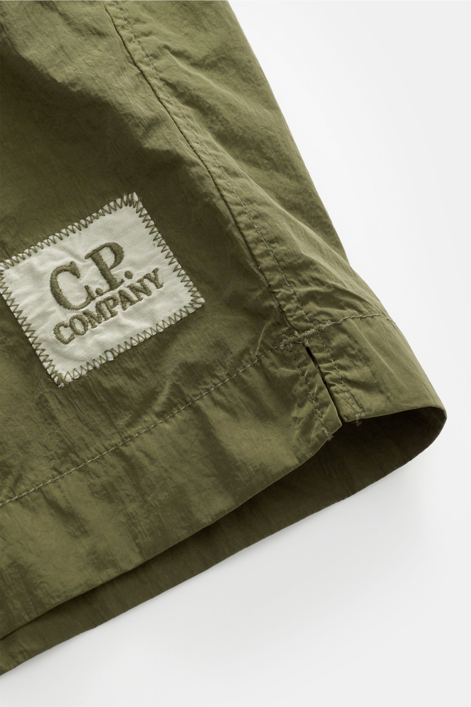 Nahaufnahme der glänzenden Oberfläche und des Logo-Patches der olivfarbenen C.P. Company Badeshorts, fotografiert von schräg oben, aus schnell trocknender Microfaser mit Regular Fit, elastischem Bund und Mesh-Innenfutter.