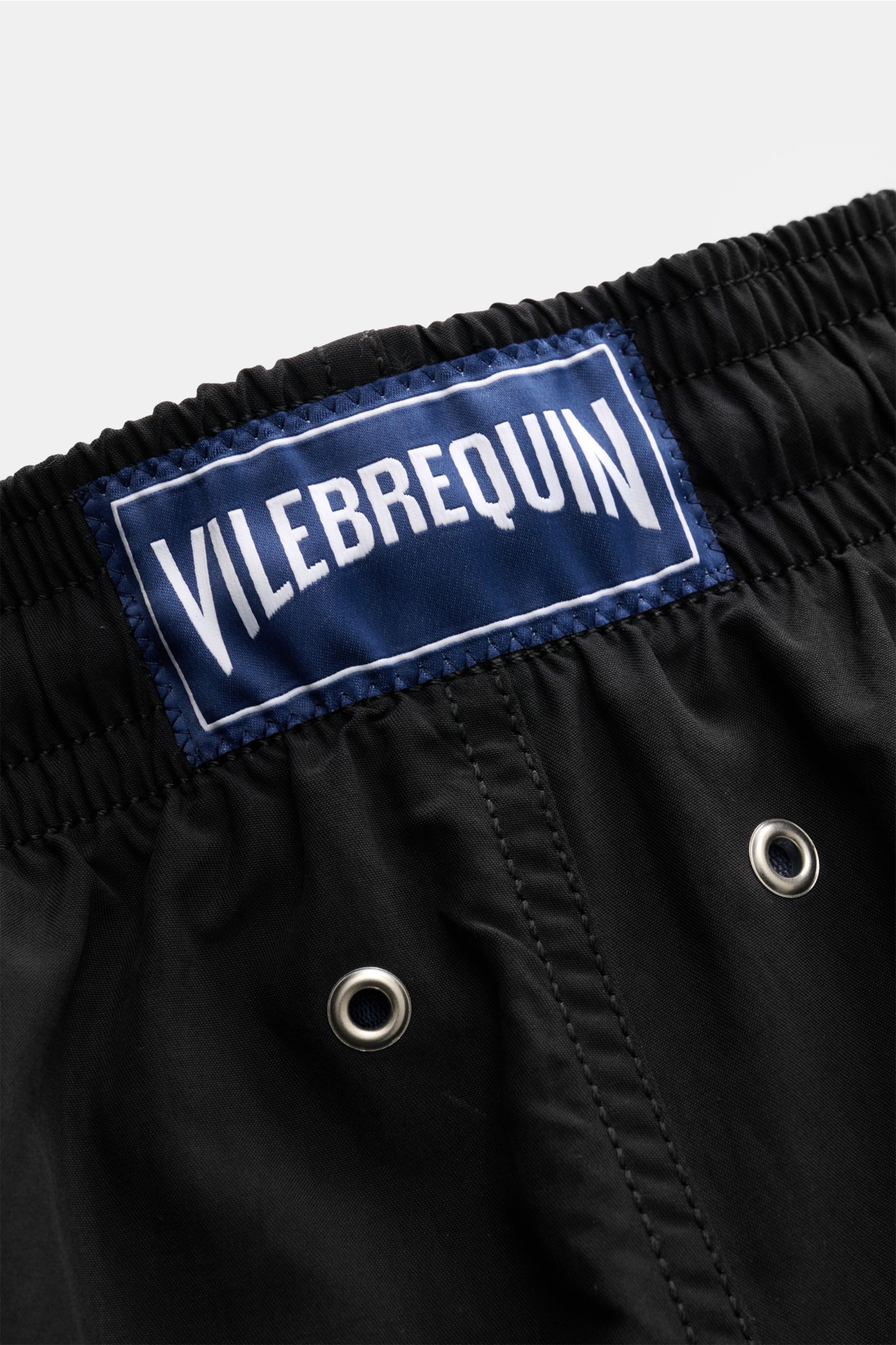 Nahaufnahme der schwarzen Vilebrequin Badeshorts 'Moorea' von hinten, zeigt elastischen Bund, Tunnelzug, Entwässerungsösen und das blaue VILEBREQUIN-Weblabel, gefertigt aus hochwertiger Microfaser mit leichtem Griff und Mesh-Innenfutter, ideal für Sommer,
