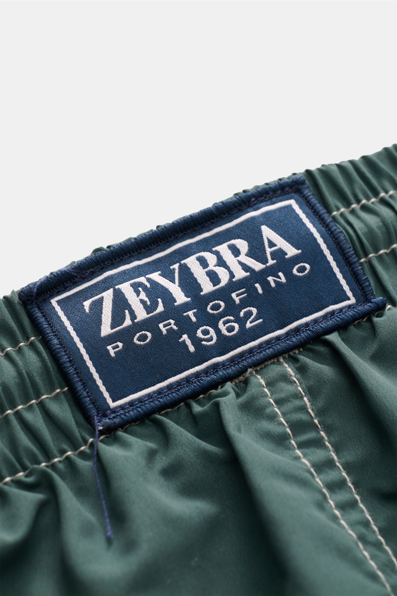 Close-up from above of the elastic waistband and ZEYBRA PORTOFINO 1962 label on Zeybra Portofino 1962 Badeshorts 'Lux Car' dunkelgrün/salbei. Sommerliches Statement: Mit ihrem markanten Print werden die Badeshorts von ZEYBRA PORTOFINO 1962 zum Blickfan