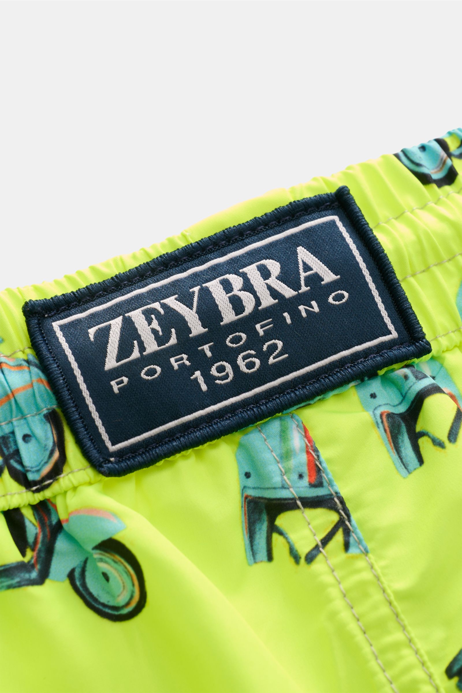 Close-up view of the waistband and label of Zeybra Portofino 1962 Badeshorts 'Scooter' neongelb/hellblau gemustert, showing the neon yellow fabric with blue scooter print and elastic waistband.

Sommerliches Statement: Mit ihrem markanten Allover-Print
