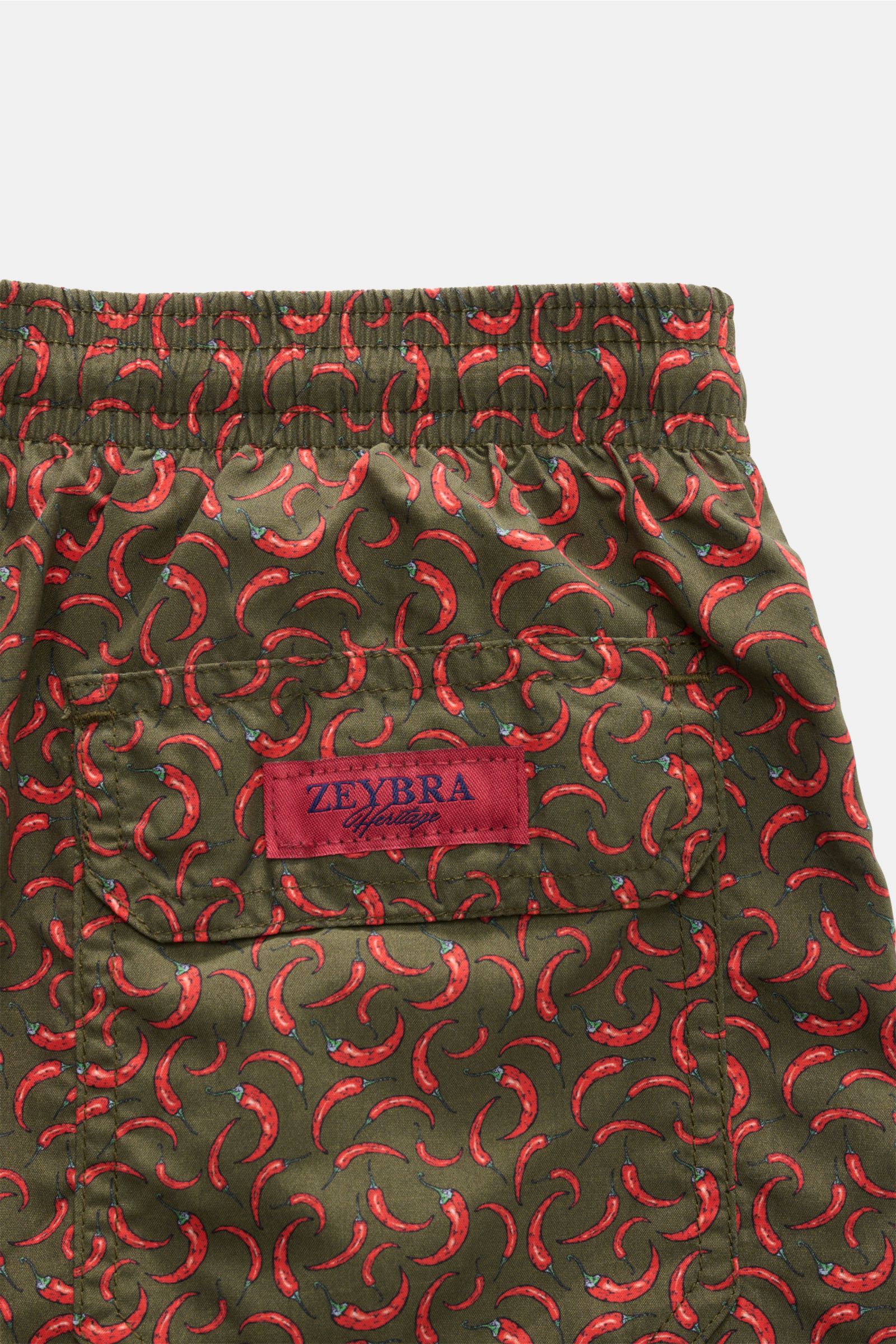 Close-up rear view of Zeybra Portofino 1962 Badeshorts 'Peperoncini' dunkelgrün/rot gemustert with elastic waistband, chili pepper print, back pocket, and red label. Eyecatcher für sonnige Tage: Die Badeshorts von ZEYBRA PORTOFINO 1962 überzeugen mit kurz