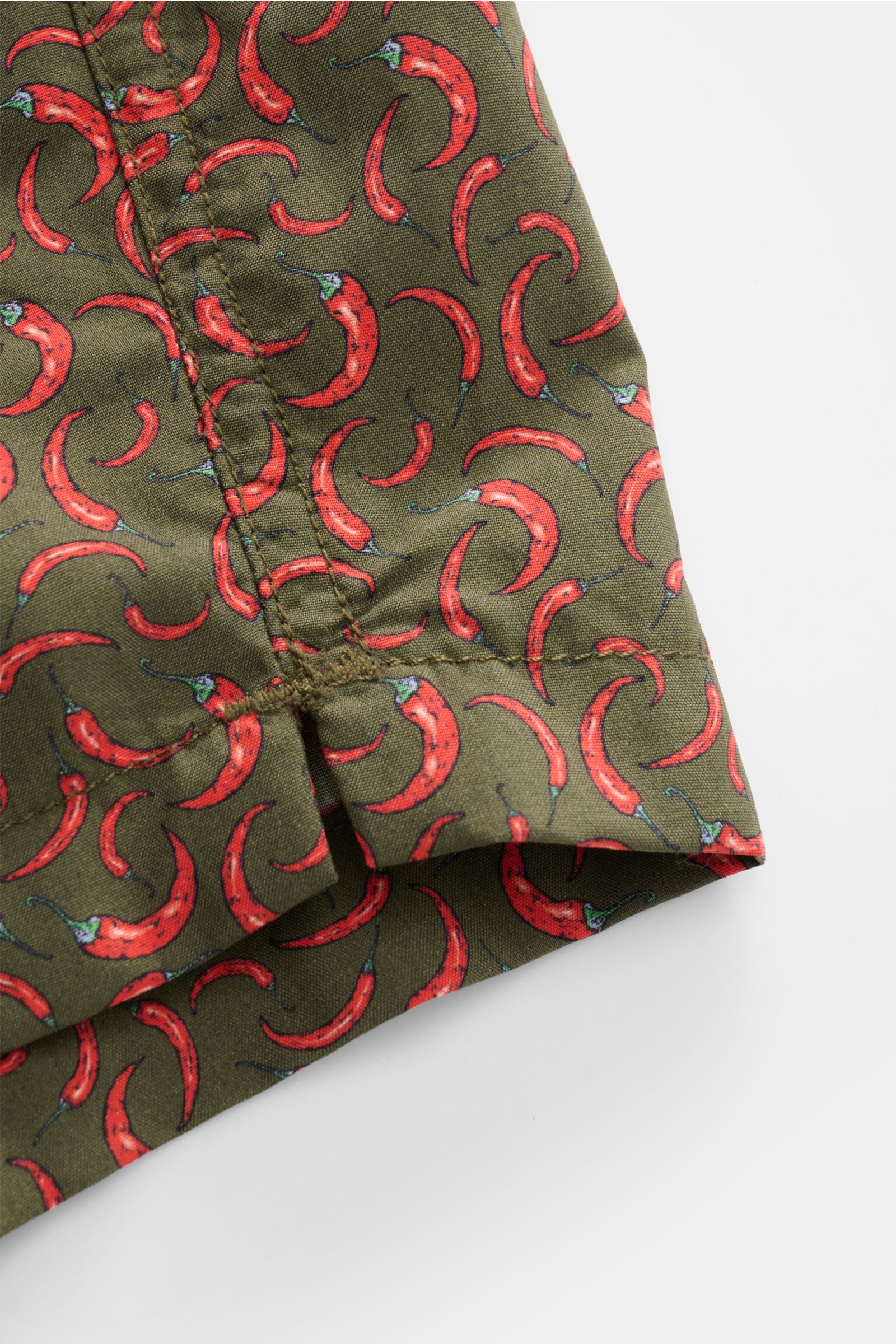 Close-up photo of the hem and seam details of the Zeybra Portofino 1962 Badeshorts 'Peperoncini' dunkelgrün/rot gemustert, showing red chili pepper print on dark green fabric, photographed from the front side.

Eyecatcher für sonnige Tage: Die Badeshor