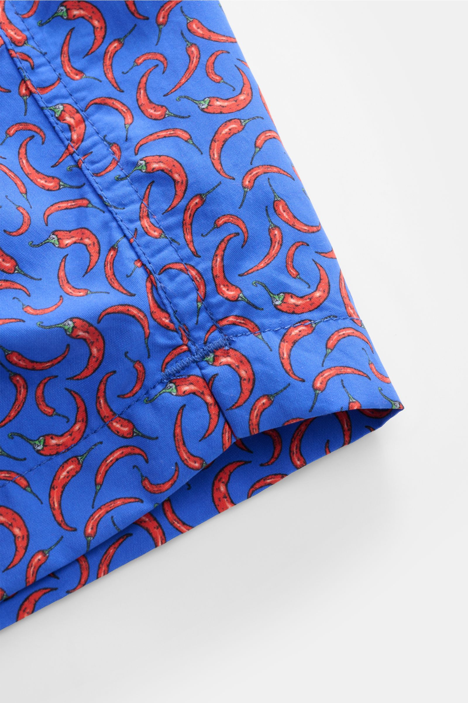 Close-up view of the hem and side seam of Zeybra Portofino 1962 Badeshorts 'Peperoncini' blau/rot gemustert, featuring a bright blue base with a red chili pepper allover print. Eyecatcher für sonnige Tage: Die Badeshorts von ZEYBRA PORTOFINO 1962 überz