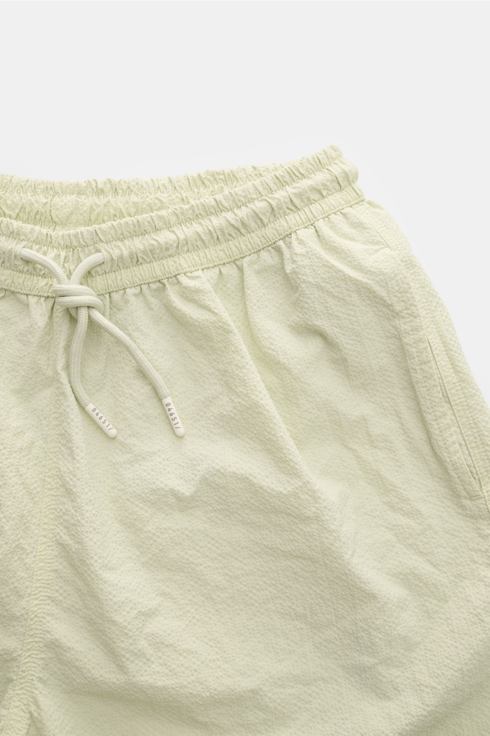 Nahaufnahme der salbeifarbenen 04651/ A trip in a bag Badeshorts 'Mono Seersucker' im Seersucker-Look mit elastischem Bund, Tunnelzug und Taschen, fotografiert von vorne.