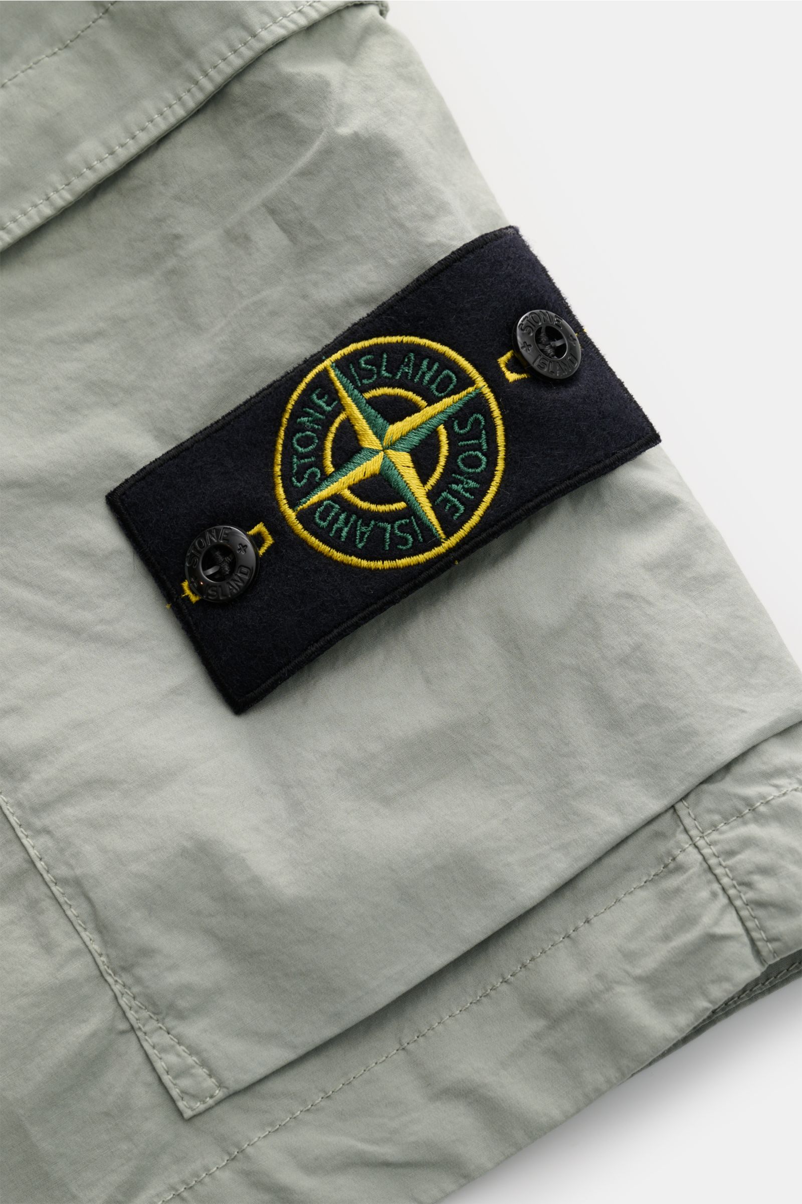 Nahaufnahme der Stone Island Cargo-Shorts salbei von schräg oben, zeigt leichte Baumwolle, Cargo-Tasche mit Knöpfen und Kompass-Badge.