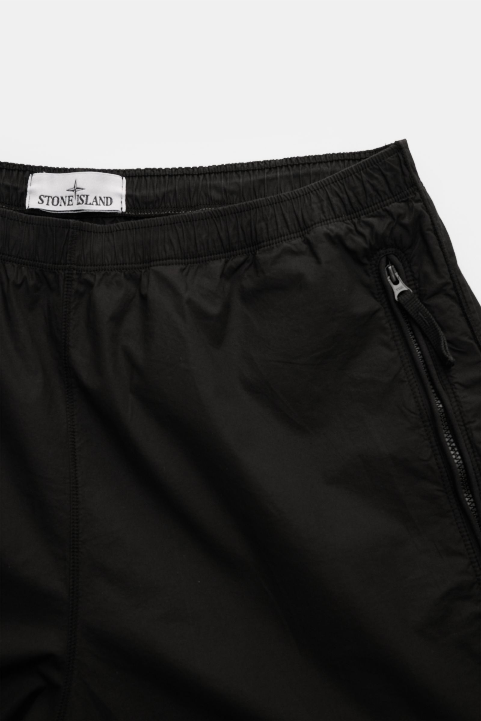 Close-up top-down view of Stone Island Shorts 'Paracadute' black, showing elasticated waistband, zipper pocket, pure cotton stretch fabric, and signature compass badge 