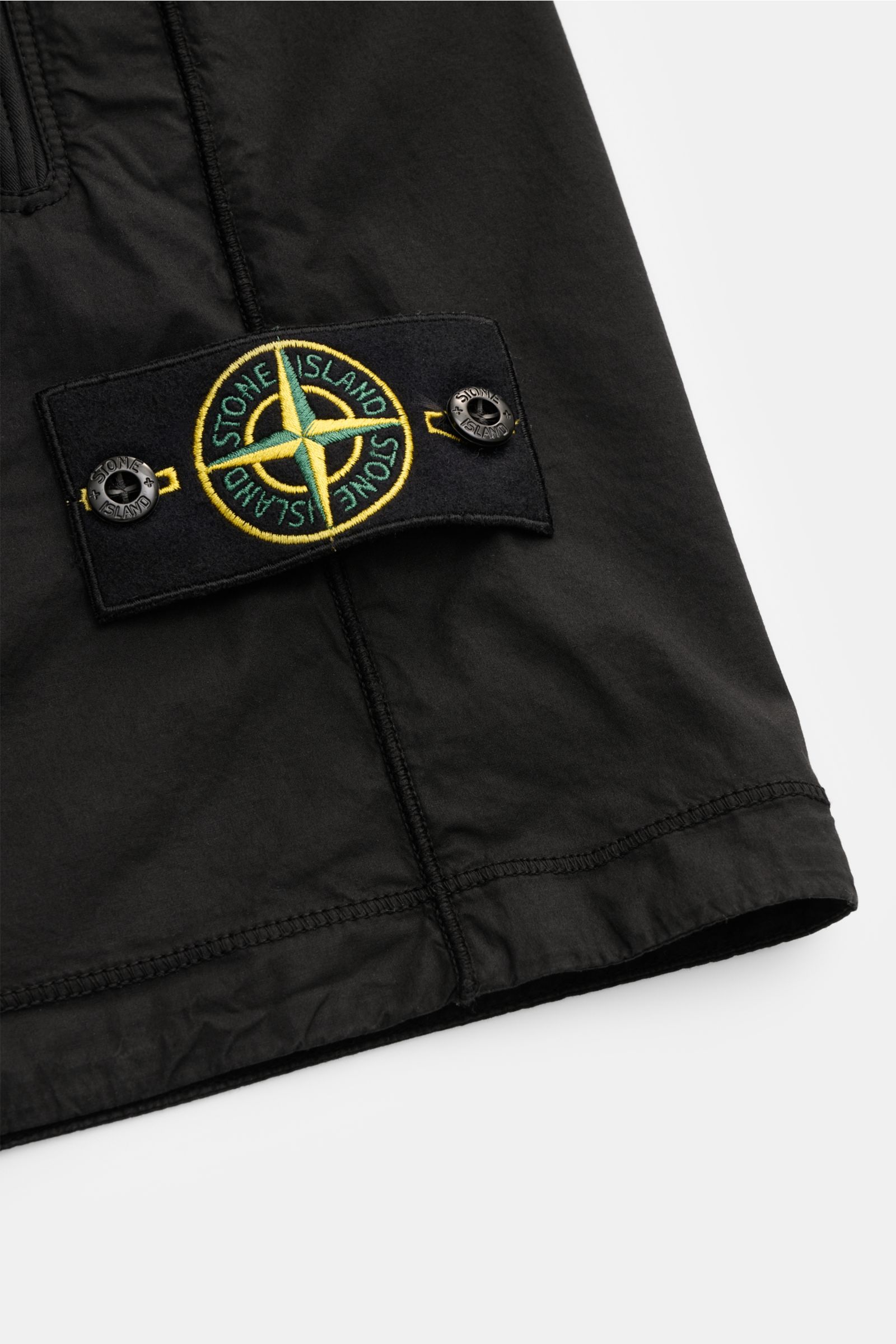Close-up view of the Stone Island Shorts 'Paracadute' black showing the distinctive compass badge with button fasteners on lightweight black cotton fabric, highlighting overlock seams and texture from a top-side perspective. Comfortable summer piece: The 
