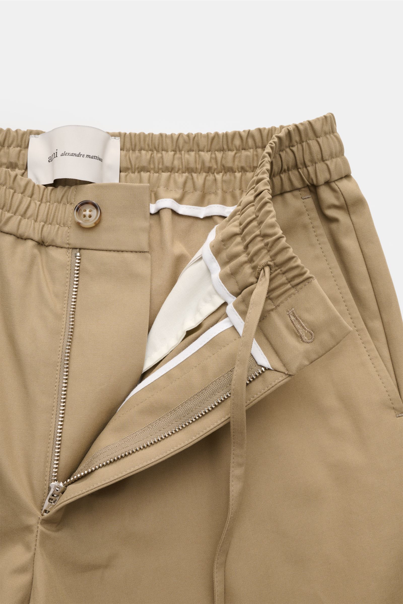 Nahaufnahme der Ami Paris Shorts taupe aus reinem Baumwoll-Twill, Regular Fit, elastischem Bund mit Tunnelzug, Reißverschluss, Knopf und französischen Taschen, Perspektive von oben auf den geöffneten Bund und Taschen.
