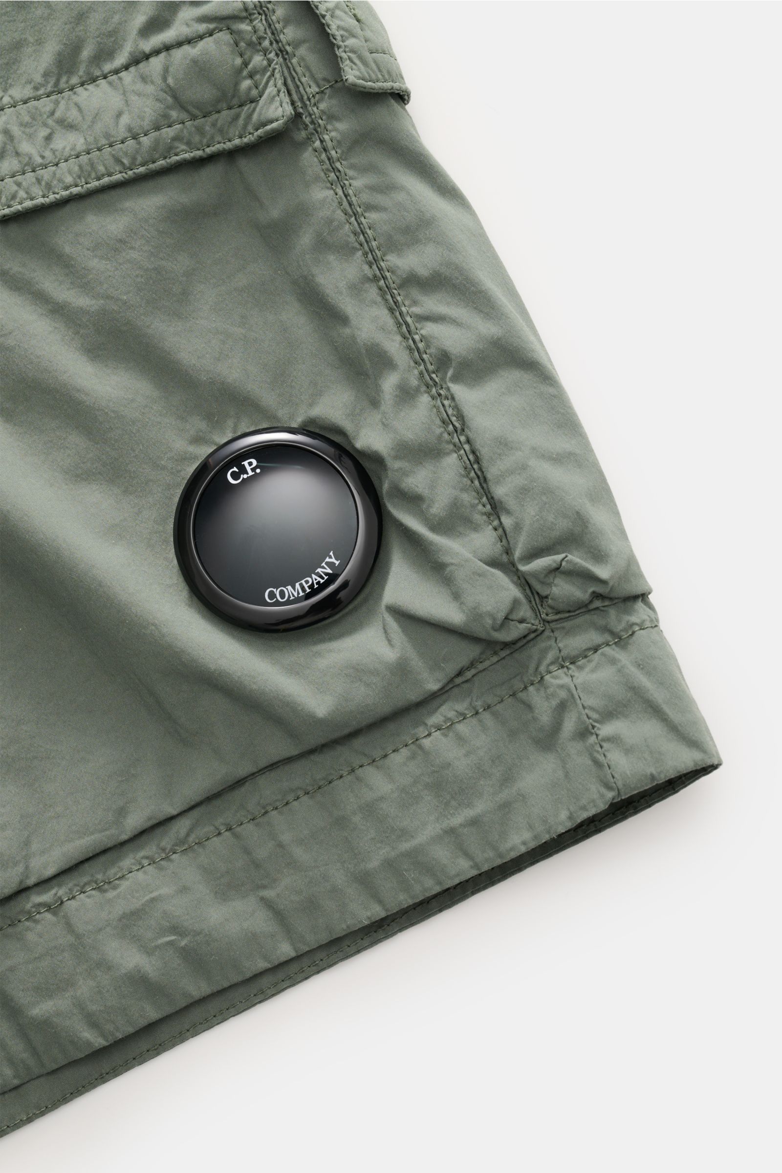 Nahaufnahme der graugrünen C.P. Company Cargo-Bermudas, schräg von oben fotografiert, mit Label-Linsendetail und Cargo-Tasche.