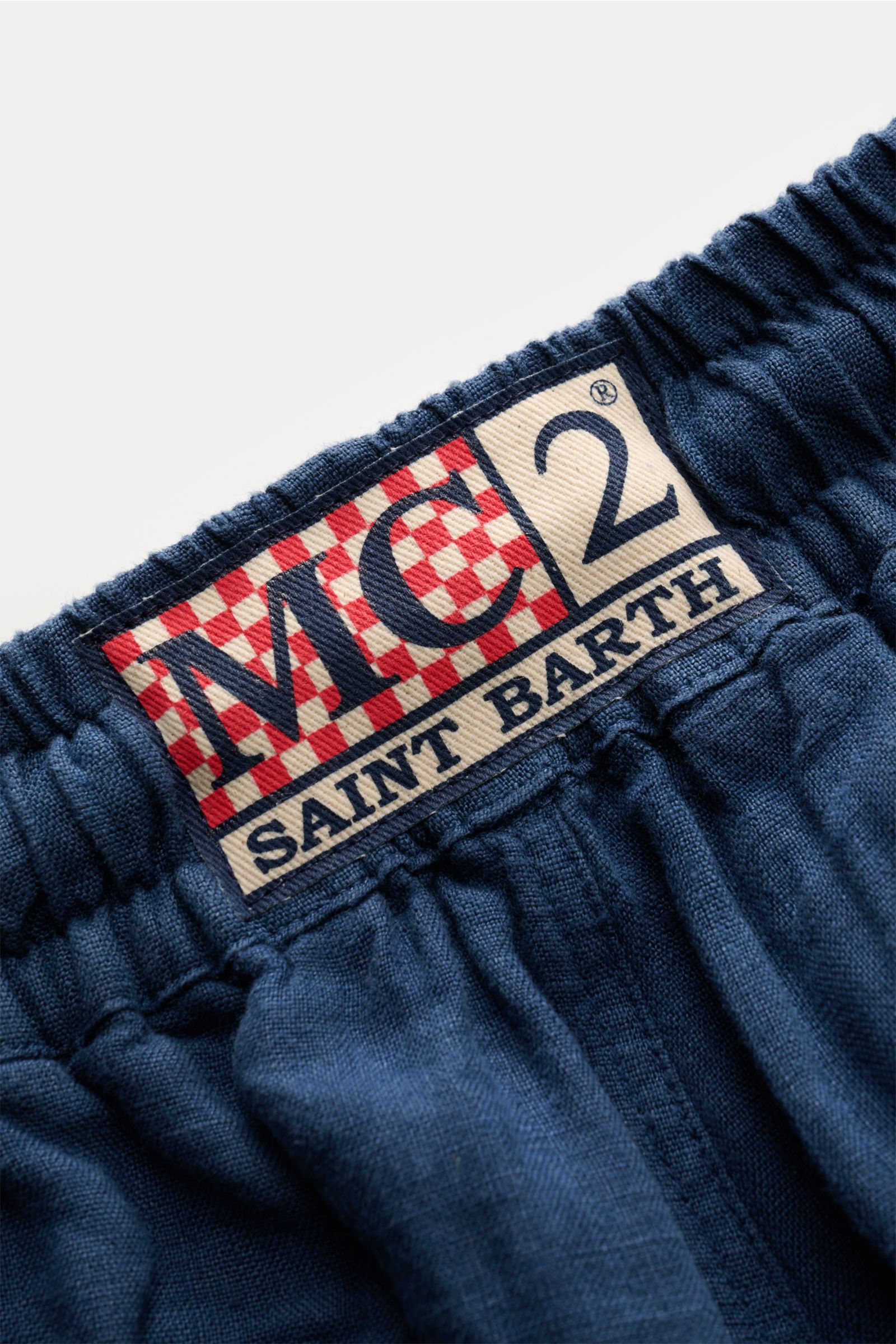 Close-up of the elastic waistband with a red and white checkered MC2 Saint Barth logo patch on the navy MC2 Saint Barth Leinen Cargo-Shorts 'Marseille' navy, showing the linen fabric texture and garment-dyed material from a top-down perspective.