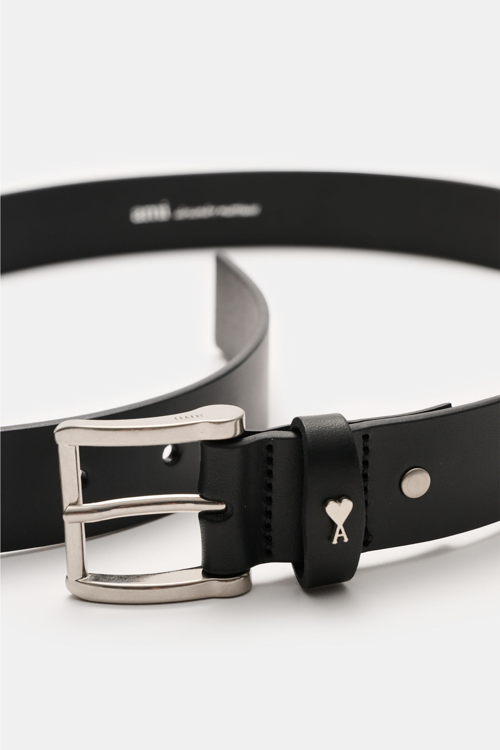 Ami Paris Gürtel 'Paris' schwarz shown close-up from a side angle, featuring a black smooth leather strap, brass prong buckle, and logo-embossed loop. Zeitloses Accessoire: Der 'Paris' Gürtel von AMI PARIS erweist sich als vielseitiges Essential für st