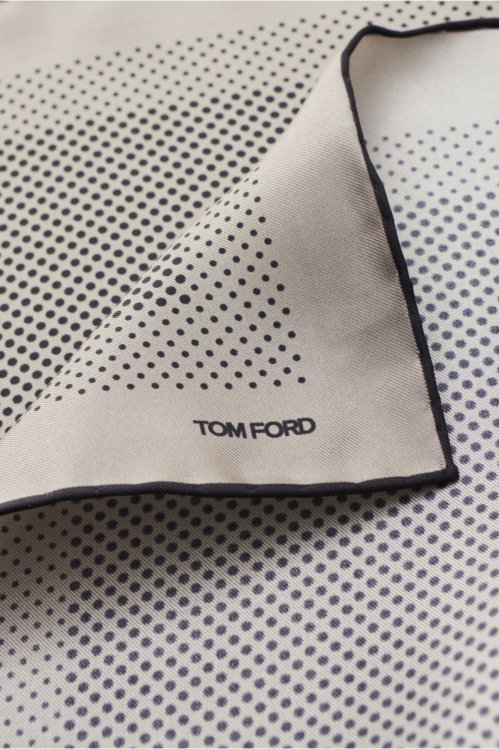 Tom Ford Einstecktuch grau/schwarz gepunktet, aus reiner Seide mit geschmeidigem Griff, modernes Punkte-Design, handrollierte Kanten, kleines TOM FORD-Logo, Made in Italy, Maße 40x40 cm, Detailaufnahme von schräg oben.