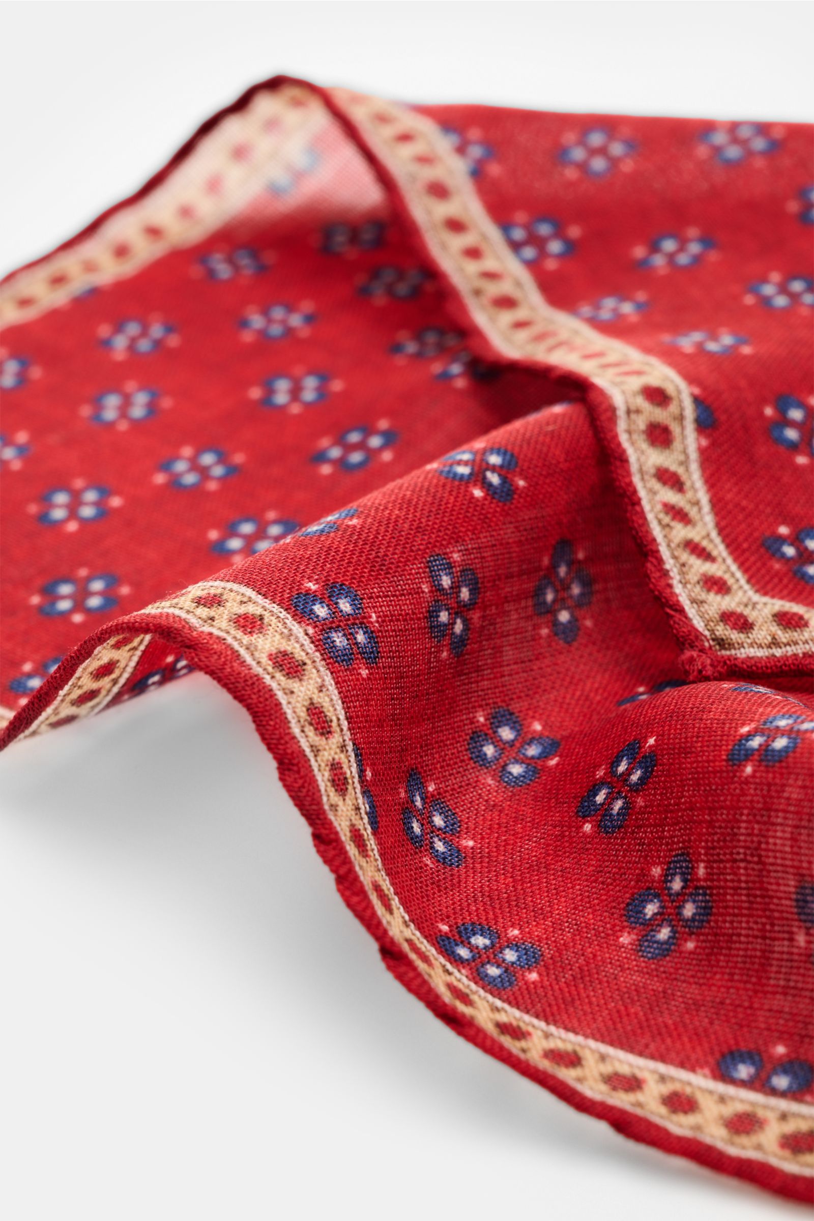 Nahaufnahme aus schräger Perspektive des Rosi Collection Einstecktuchs 'Ted' rot meliert/dunkelblau/beige gemustert aus reiner Wolle mit floralem Print, Borte und rollierten Kanten, Maße ca. 30x30 cm, Made in Italy.