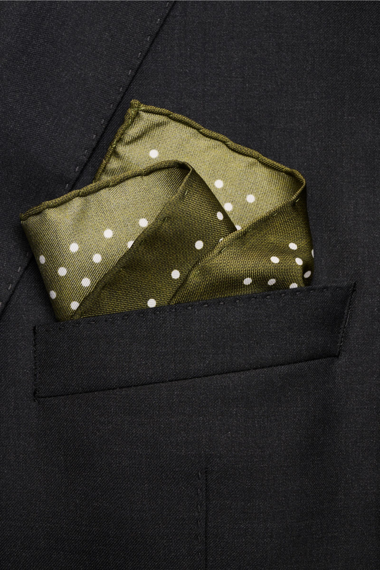 Rosi Collection Silk pocket square olive with polka dots folded in a black jacket pocket, photographed from a top front angle. Pure silk, smooth touch, graphic polka dot pattern, hand-rolled edges, made in Italy, dimensions approx. 31 cm x 31 cm.