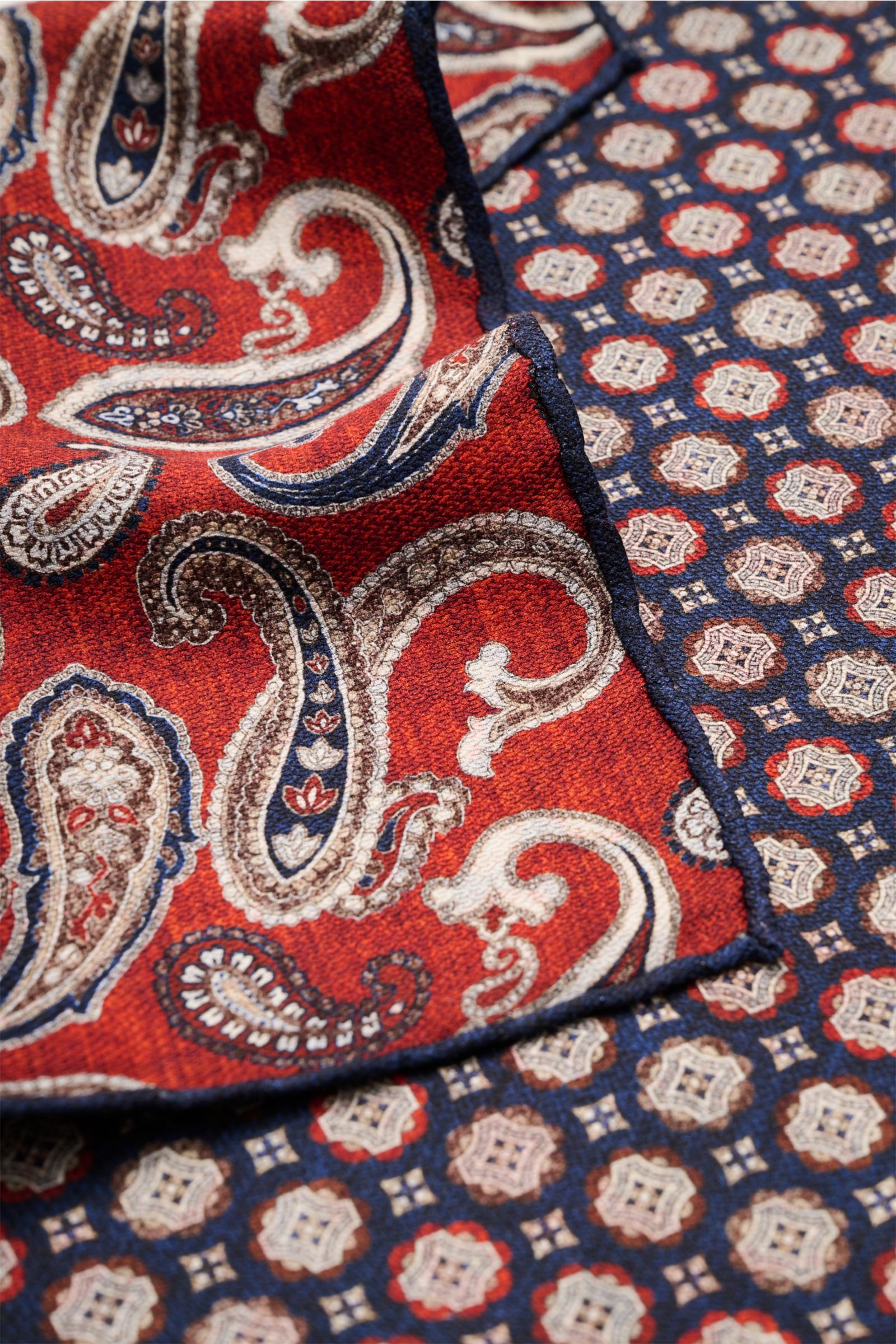 Close-up image of the Rosi Collection Silk pocket square 'Easy' navy/dark brown/rust brown patterned, photographed from a top angle showing the elegant medallion print on the front and the paisley pattern on the back with hand-rolled edges. Pure silk with