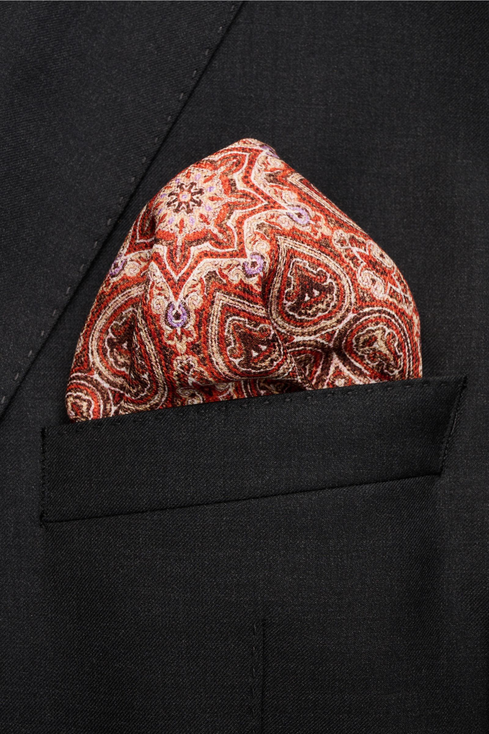 Rosi Collection Einstecktuch 'Easy' rot/orange/beige gemustert mit modernem Paisley-Muster aus reiner Seide, rollierte Kanten, Vorder- und Rückseite unterschiedlich, in schwarzem Sakko aus schräger Perspektive.