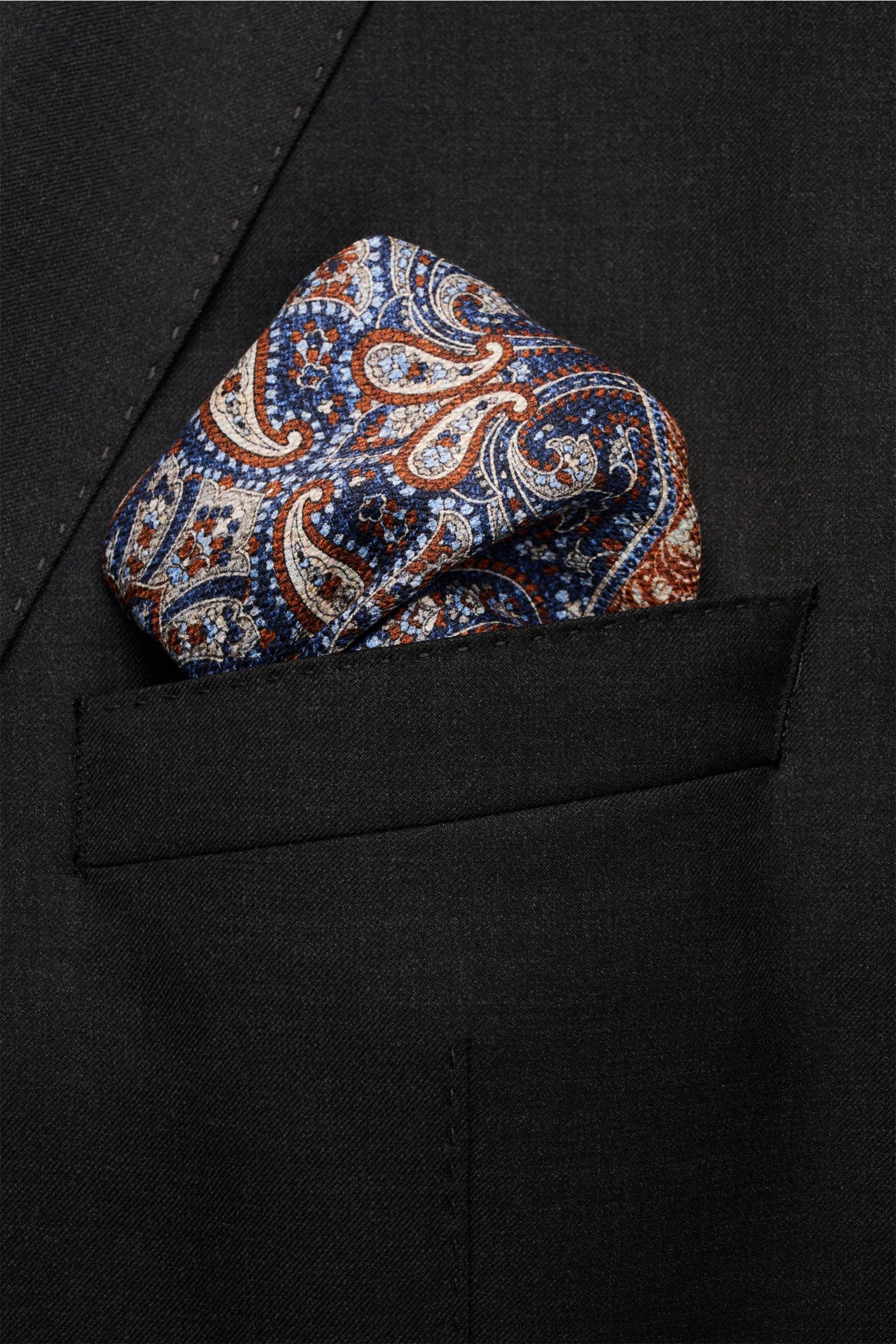 Rosi Collection Seiden-Einstecktuch 'Easy' graublau/blau/braun gemustert, Paisley- und Kachel-Muster, reine Seide, 31x31 cm, handrollierte Kanten, Rückseite Medaillon-Print, softer Griff, Made in Italy, seitliche Nahaufnahme in schwarzer Brusttasche.