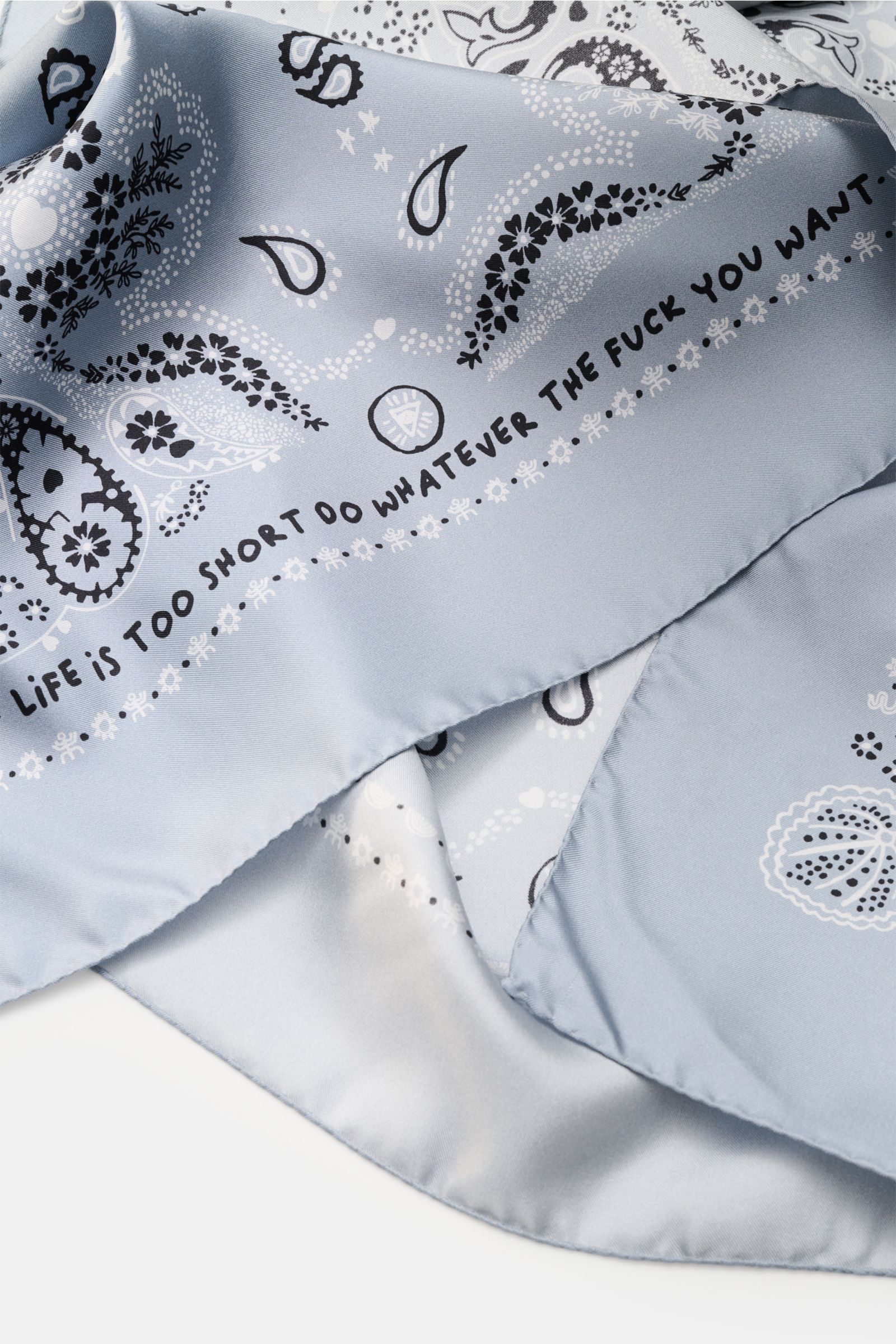 Close-up from a top angle of the Ace Denim Silk scarf smoky blue/black/white patterned with paisley print and bold text on smooth pure silk.

Additional description:  
Eye-catching for the warm season: The ACE DENIM silk scarf in a bandana design is a 