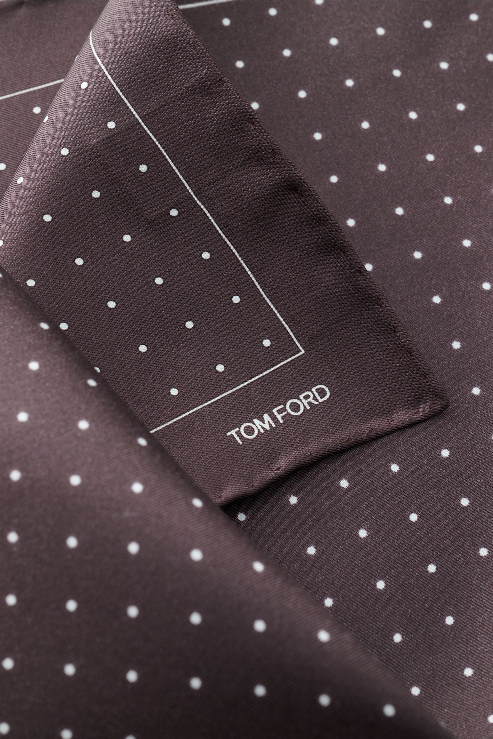 Close-up top-down view of Tom Ford Seiden-Einstecktuch dunkelbraun/weiß gepunktet with delicate white dots, rollierte Kanten, and small TOM FORD print. Dieses Einstecktuch von TOM FORD aus reiner Seide setzt mit seinem dezenten Punktmuster und den roll
