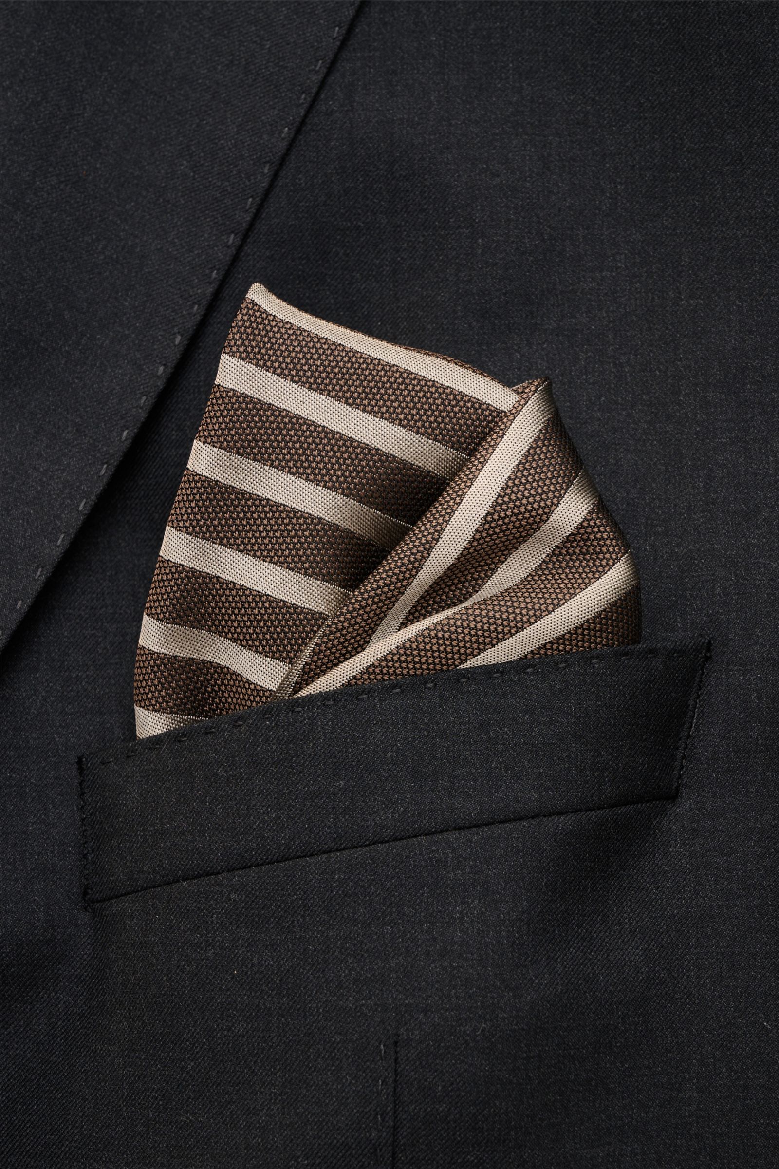 Close-up front view of a Brunello Cucinelli Seiden-Einstecktuch dunkelbraun/taupe gestreift pocket square in a black jacket pocket, showing its striped pattern, pure silk, smooth texture, hand-rolled edges, made in Italy, size approx. 35 cm x 35 cm.