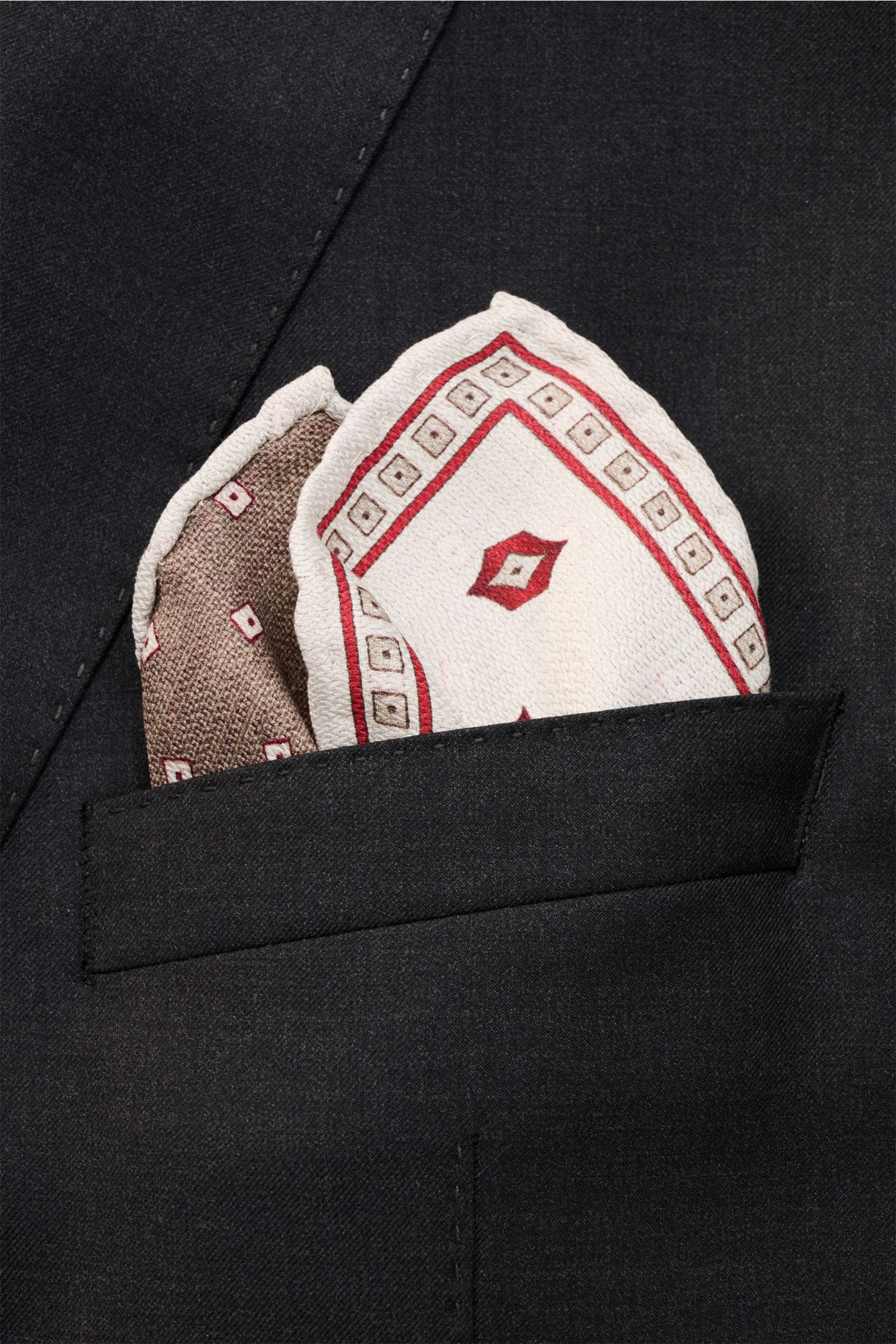 Close-up from the front of the Brunello Cucinelli Seiden-Einstecktuch braun/rot/off-white gemustert folded in a black jacket pocket; reine Seide, geschmeidiger Griff, handrollierte Kanten, Made in Italy, Maße: ca. 35 cm x 35 cm.