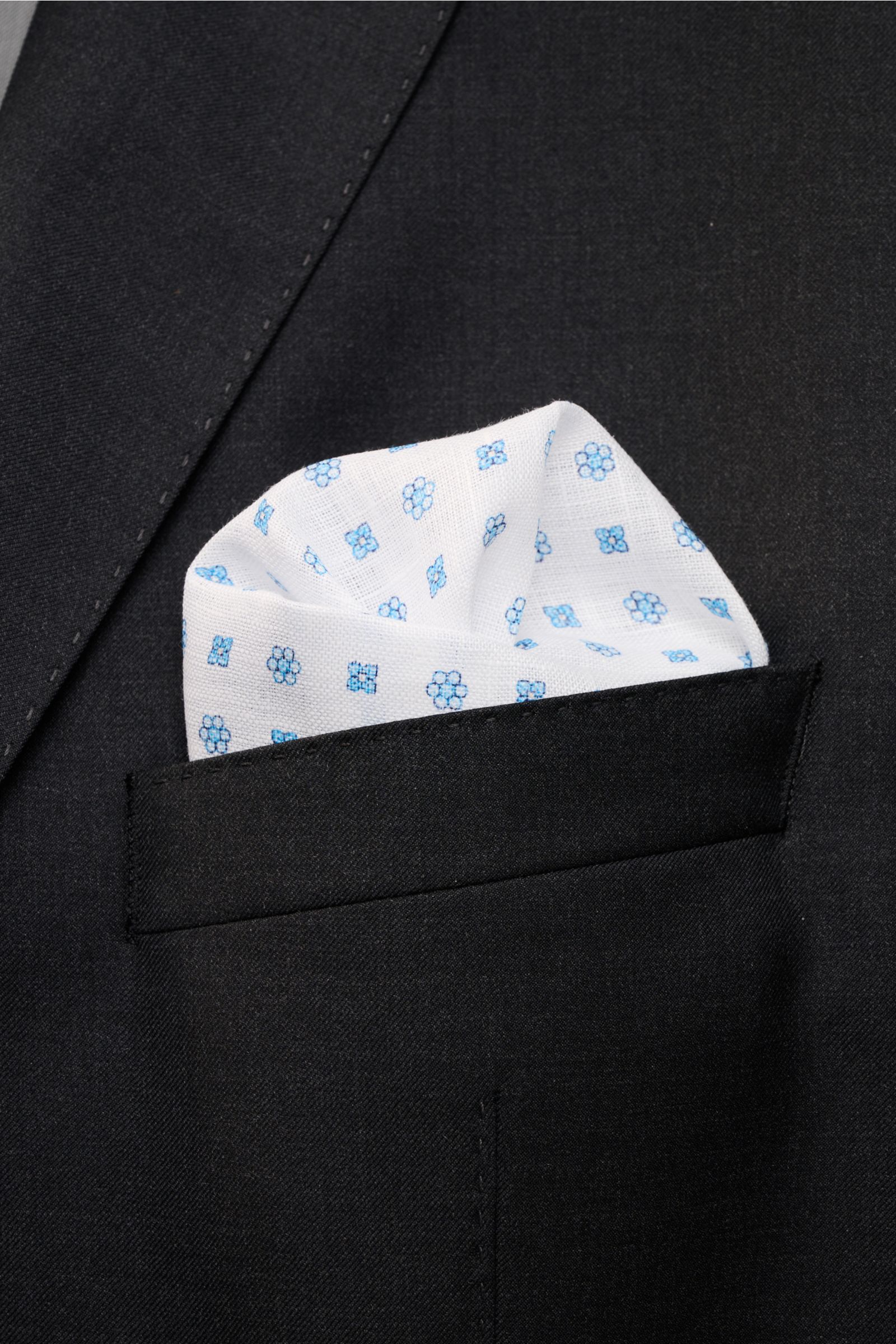 Rosi Collection Linen pocket square 'Fox' white/light blue patterned, soft pure linen with floral print and rolled edges, made in Italy, 31.5x31.5 cm, shown folded in a black jacket pocket from a close-up front angle.