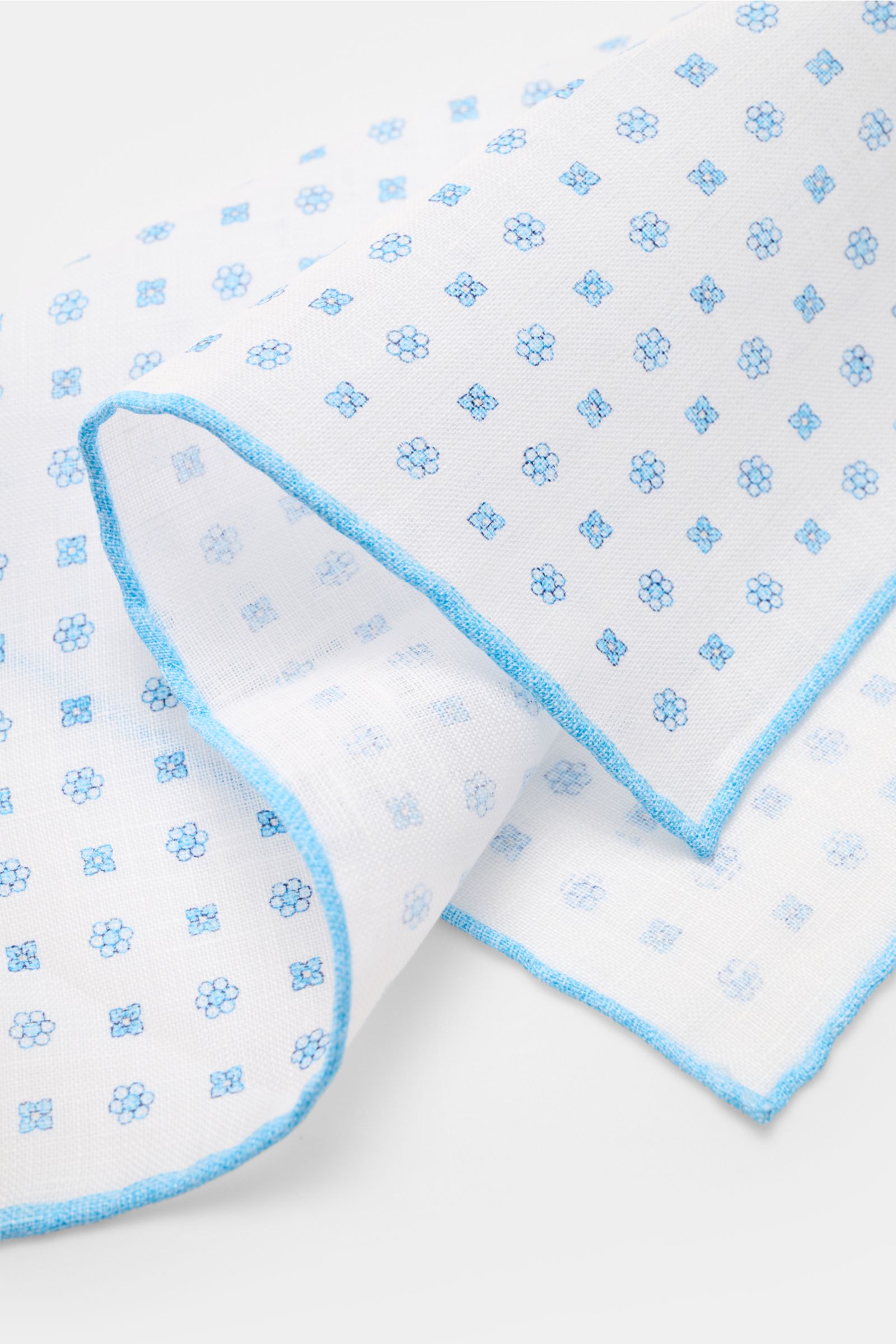 Rosi Collection Linen pocket square 'Fox' white/light blue patterned, pure linen with soft feel, floral print, rolled edges, made in Italy, 31.5x31.5 cm, shown close-up from a side angle.