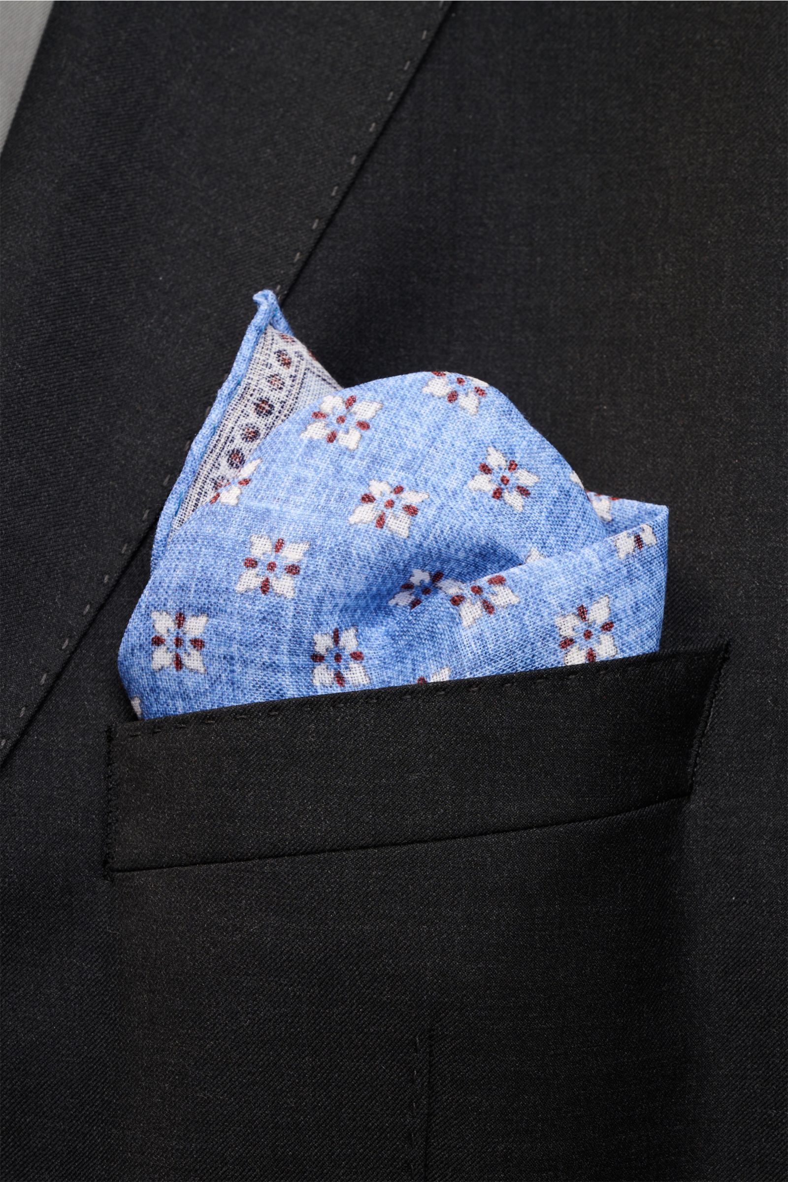 Close-up front view of the Rosi Collection Linen-pocket square 'Fox' blue/red-brown/off-white patterned in a black suit pocket, featuring pure linen, soft feel, floral print with edging, rolled edges, made in Italy, dimensions 31 cm x 31 cm.