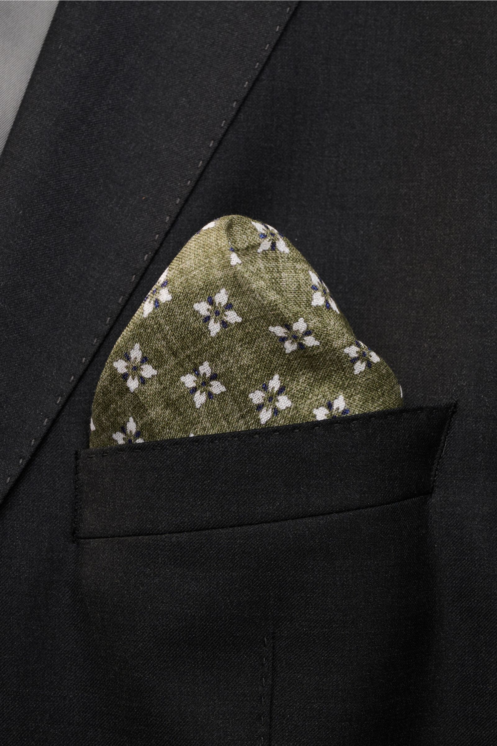 Close-up front view of the Rosi Collection Linen pocket square 'Fox' olive/dark blue/white patterned, pure linen, soft feel, floral print with edging, rolled edges, made in Italy, dimensions 31 cm x 31 cm, tucked neatly into a black suit jacket pocket.