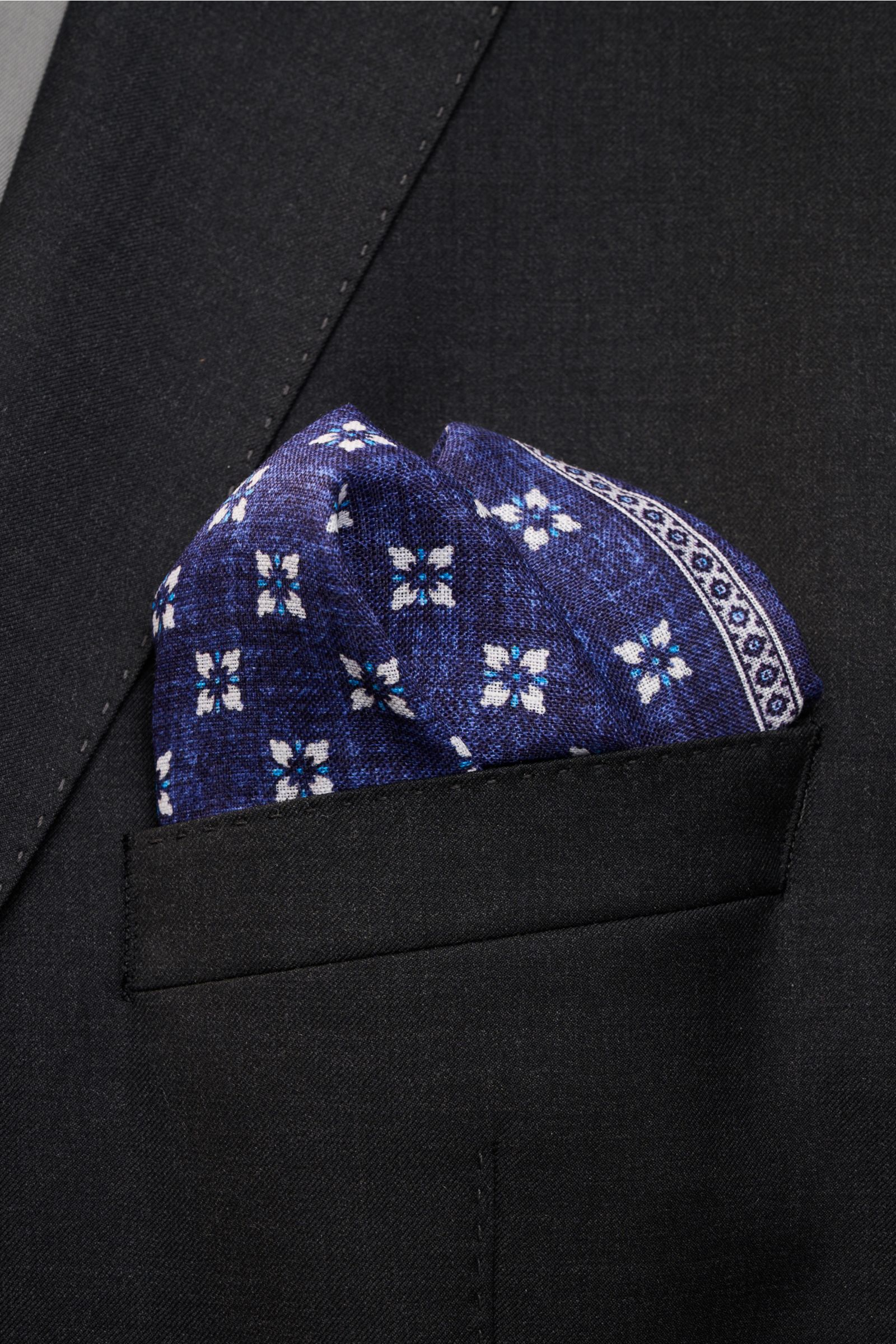Rosi Collection Linen pocket square 'Fox' dark blue/light blue/white patterned in a black jacket pocket, close-up front angle view, pure linen, soft feel, floral print with edging, rolled edges, made in Italy, dimensions 31 cm x 31 cm.