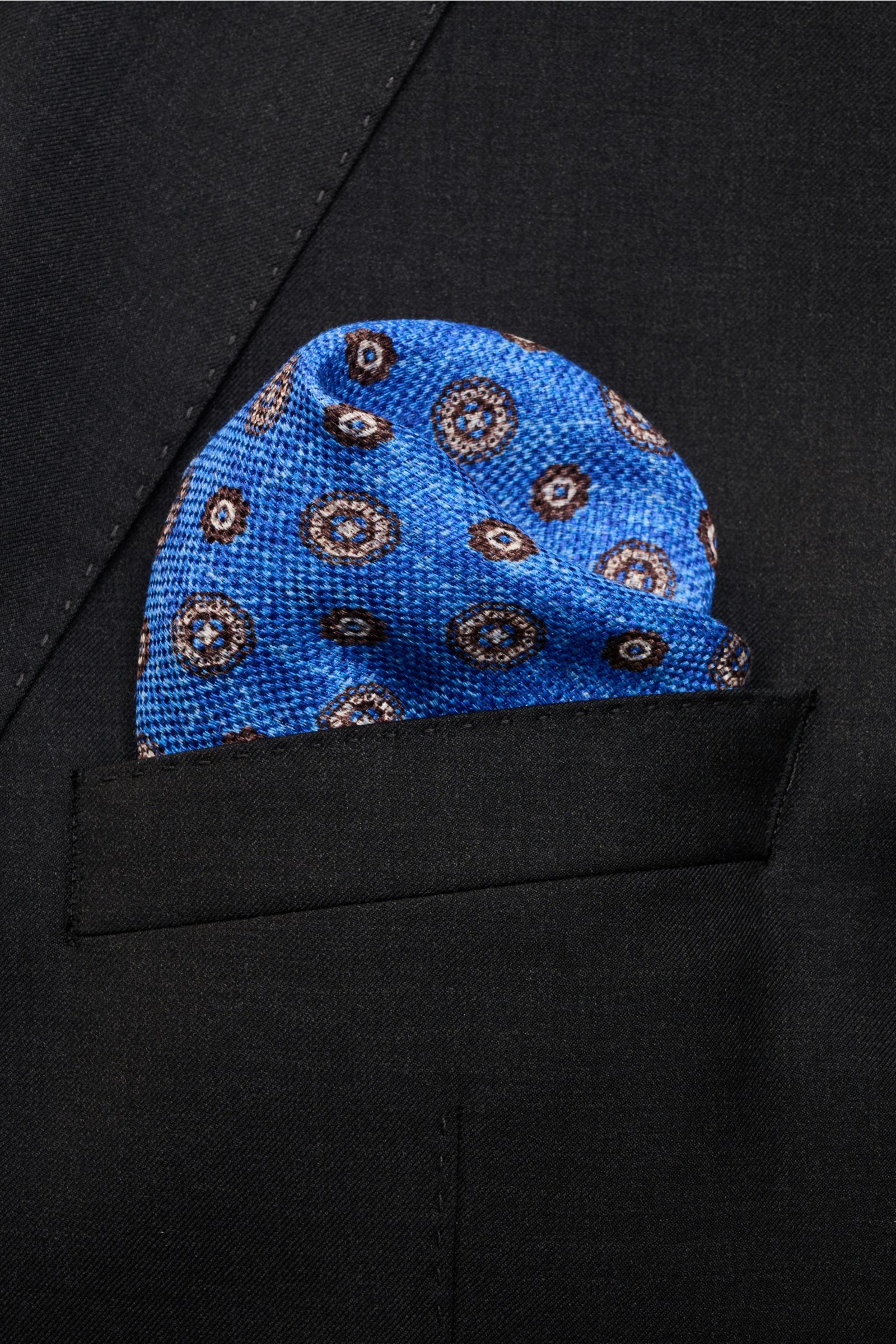 Nahaufnahme des Rosi Collection Einstecktuchs 'Joy' blau/weiß/dunkelbraun gemustert mit floralen Retro-Motiven, aus Seide und Leinen, in der Brusttasche eines schwarzen Sakkos, von vorne fotografiert.