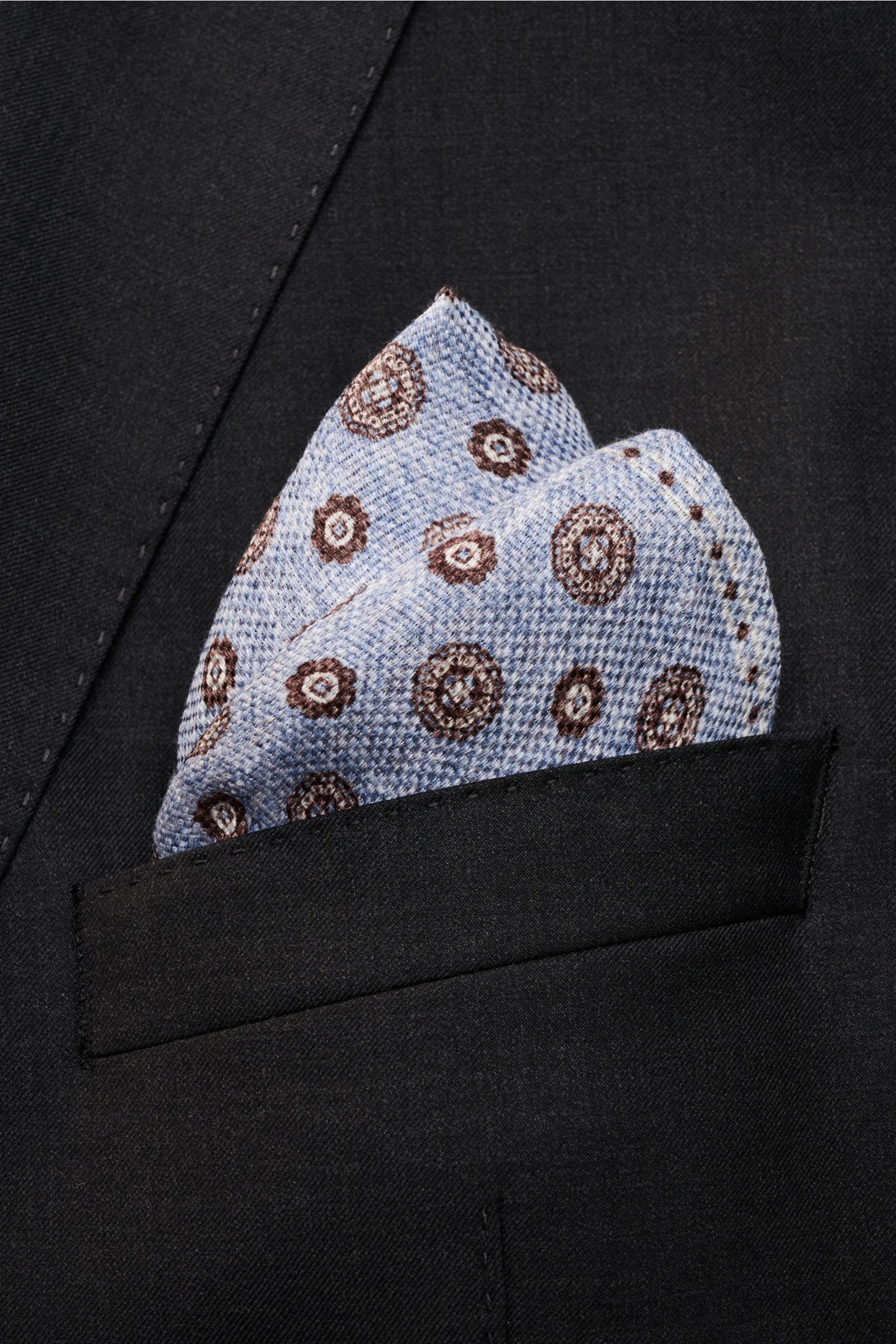 Nahaufnahme aus schräger Perspektive des Rosi Collection Einstecktuchs 'Joy' rauchblau/weiß/dunkelbraun gemustert im Sakko, florales Retro-Muster, Seide-Leinen-Mix, rollierte Kanten, Vorder- und Rückseite unterschiedlich, ca. 31x31 cm, Made in Italy.