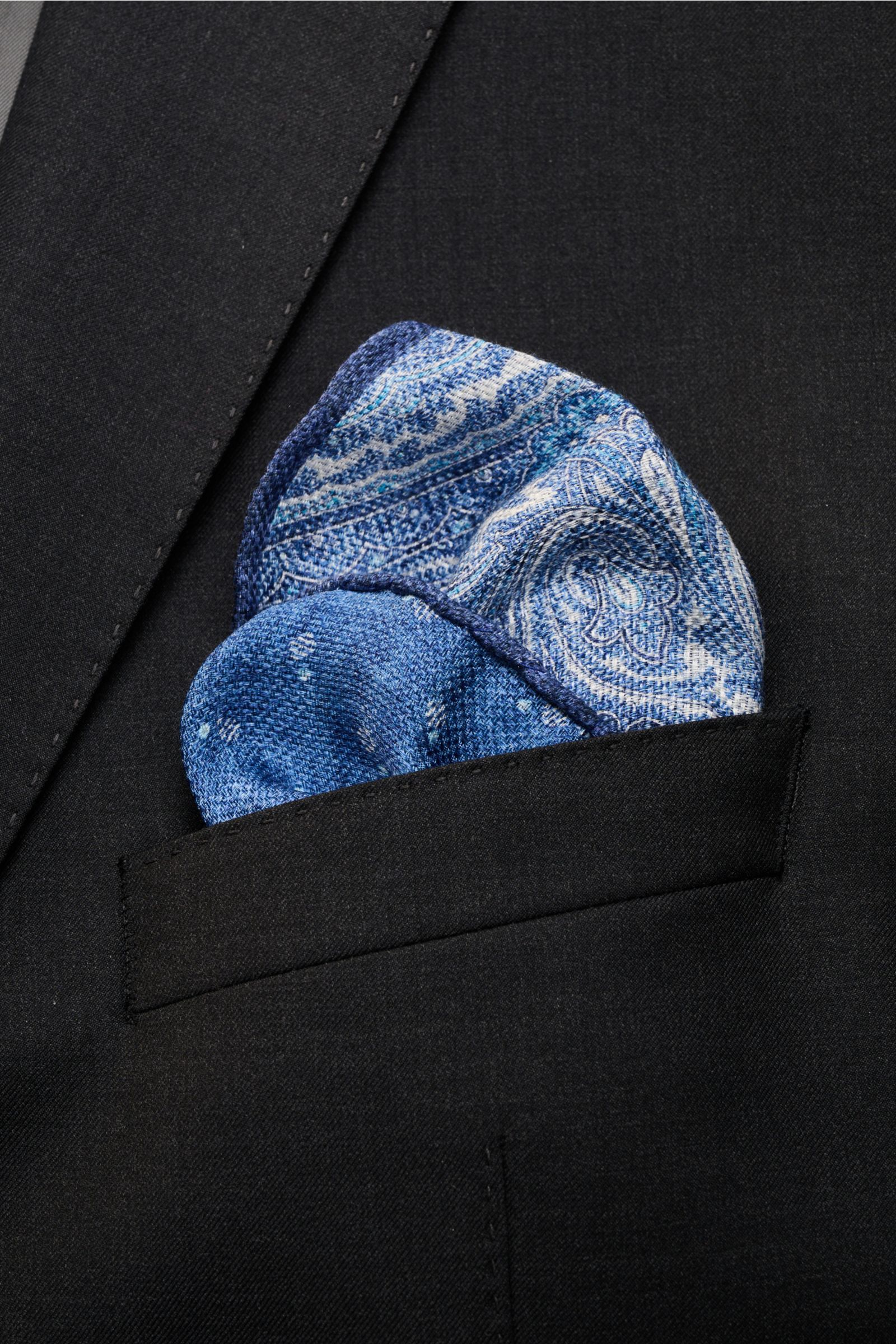 Close-up from above of the Rosi Collection Einstecktuch 'Joy' dunkelblau/weiß/hellblau gemustert pocket square with a modern paisley pattern tucked into a black jacket pocket.

Eleganter Eyecatcher: Mit seinem modernen Paisley-Muster wird das Einsteckt