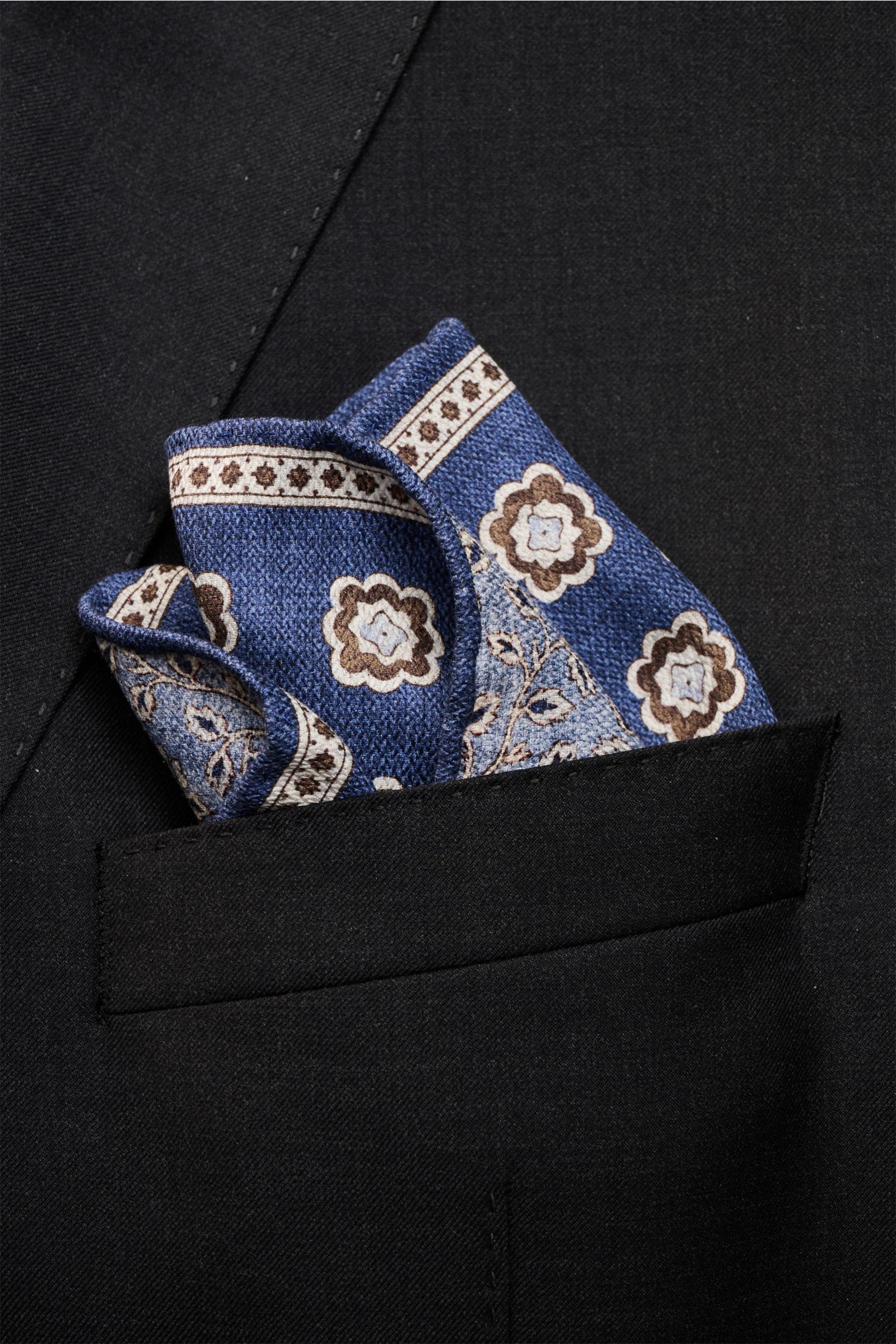 Nahaufnahme aus schräger Perspektive eines Rosi Collection Seiden-Einstecktuchs 'Easy' dunkelblau/braun/hellblau gemustert in der Brusttasche eines schwarzen Sakkos, reine Seide, softer Griff, eleganter Medaillon-Print mit Borte, florale Rückseite, rollie