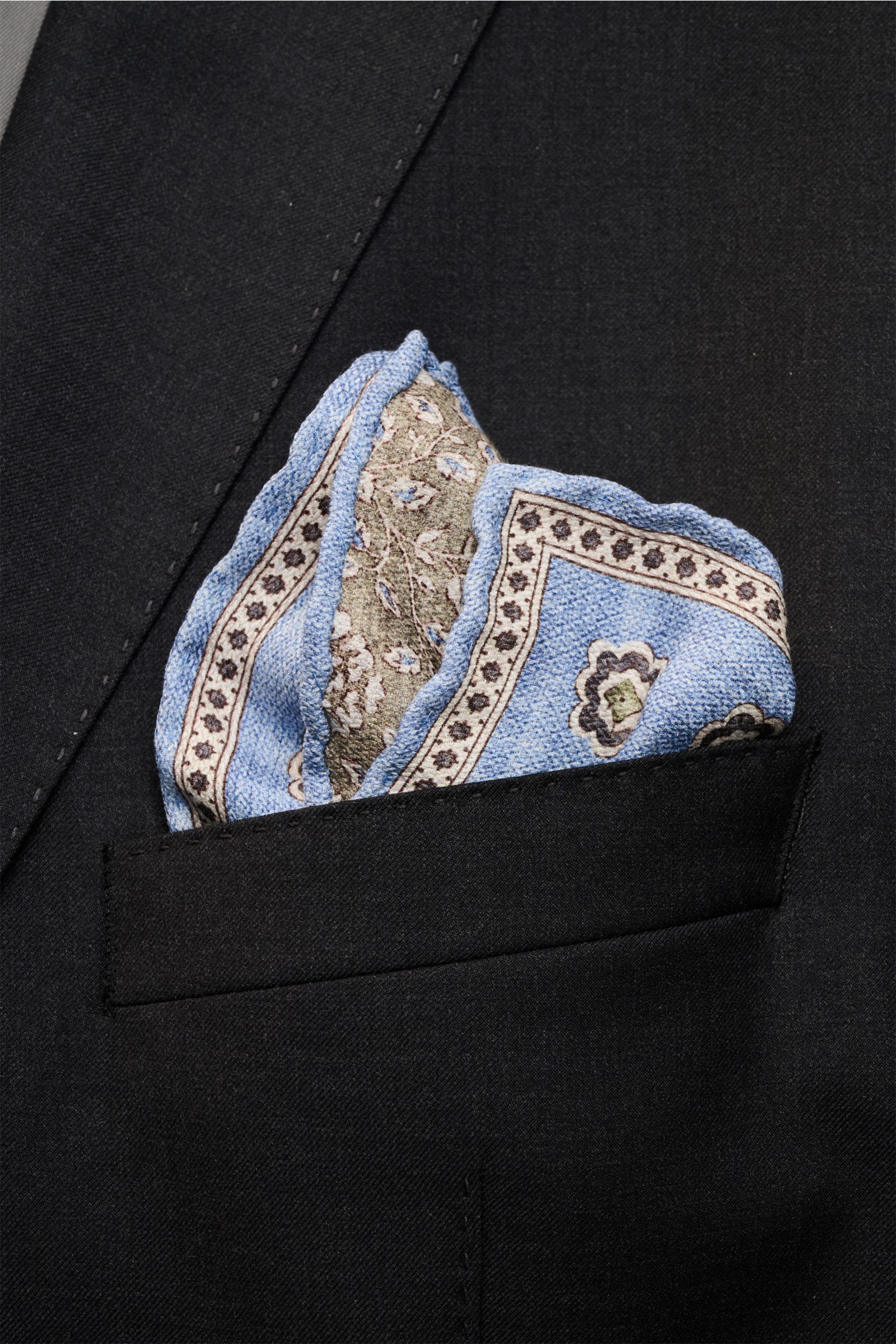 Rosi Collection Seiden-Einstecktuch 'Easy' hellblau/oliv/creme gemustert, reine Seide, softer Griff, Medaillon-Print mit Borte, florale Rückseite, rollierte Kanten, Made in Italy, ca. 31x31 cm, steckend in schwarzer Jackettasche, Frontalaufnahme.