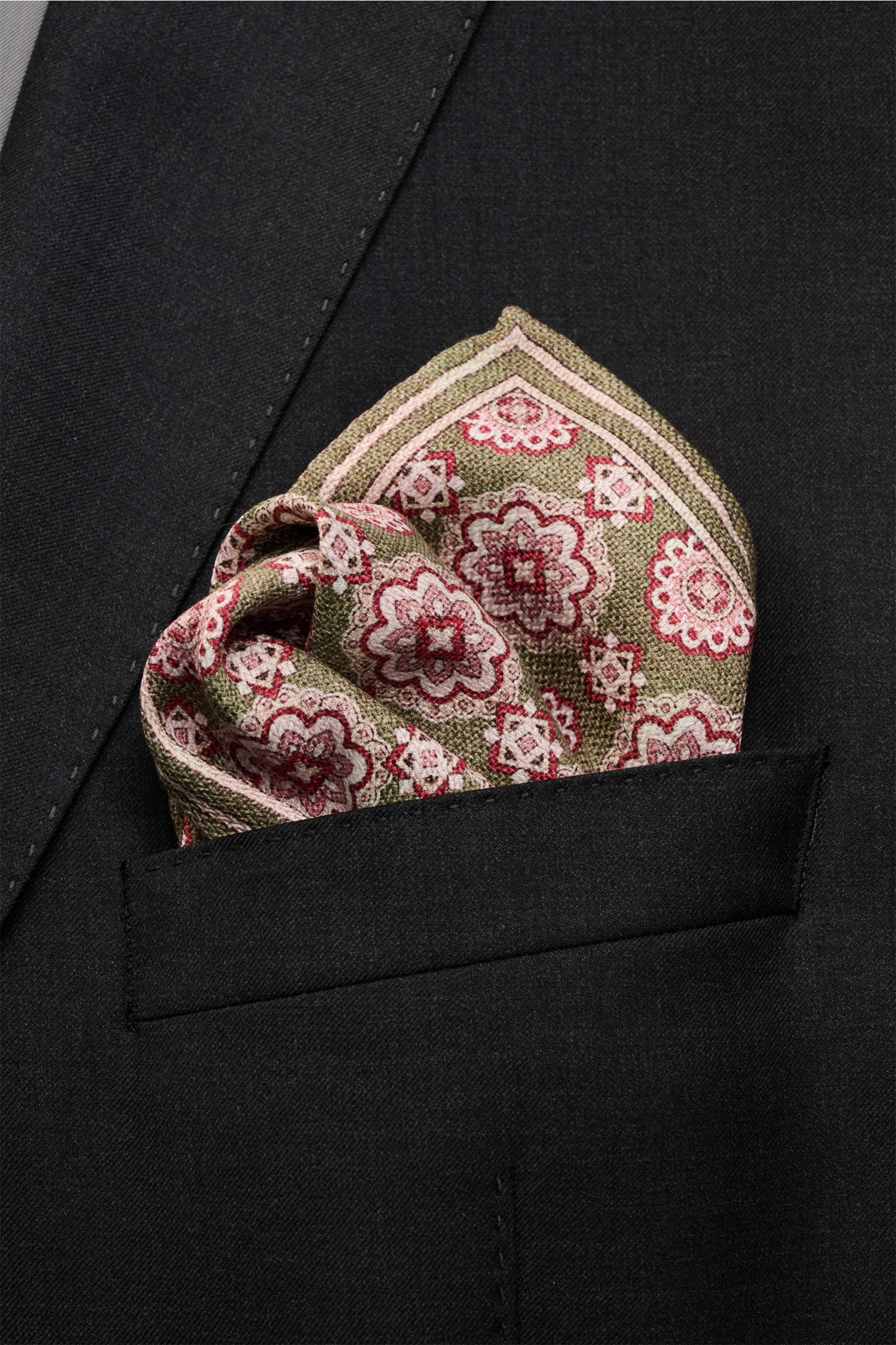 Close-up front view of a black suit jacket pocket featuring the Rosi Collection Seiden-Einstecktuch 'Easy' oliv/rot/rosé gemustert, a pocket square made of reine Seide with a softer, geschmeidiger Griff, elegant Medaillon-Print mit Borte, contrasting Rück